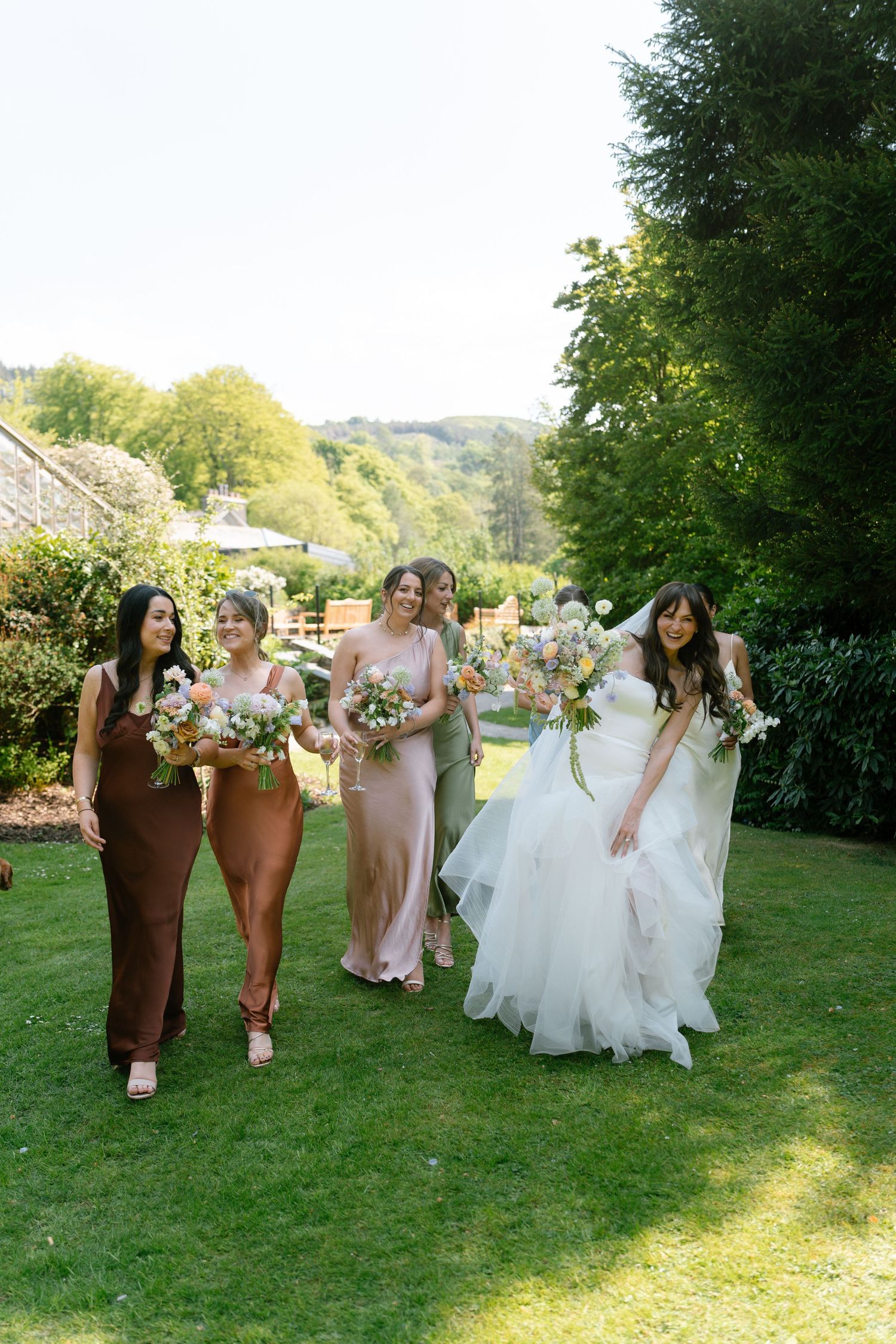 Wedding party members in earth-toned dresses walk together holding bouquets on a grassy hill.