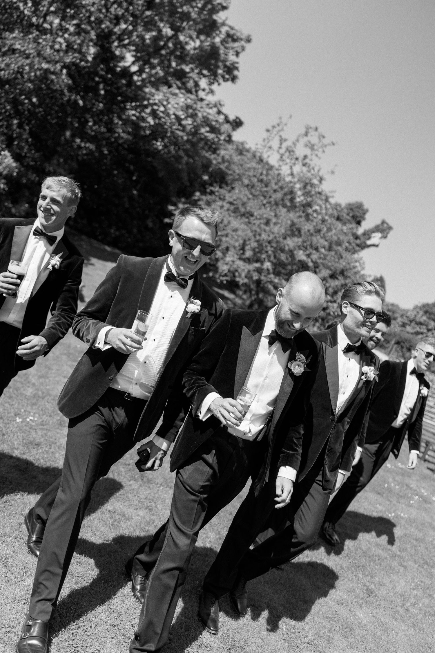 Wedding party members in formal attire pose together while holding beverages outdoors.