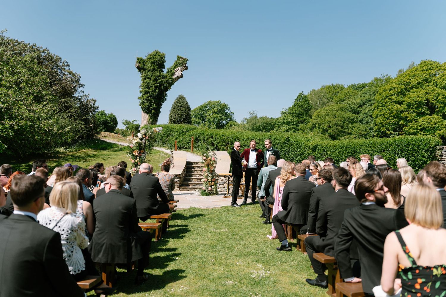 Panoramic view of wedding guests seated on wooden benches during an outdoor ceremony.