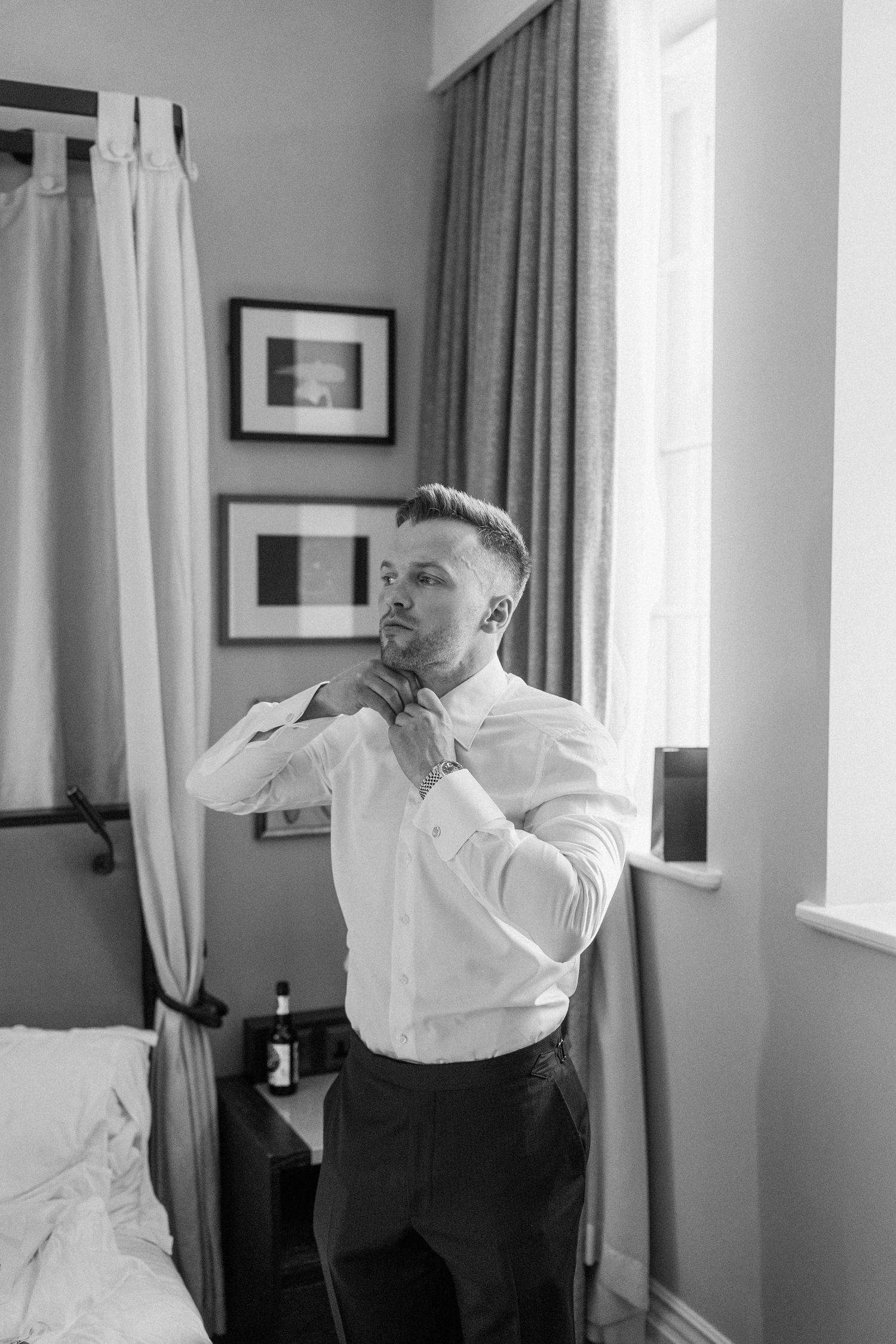Black and white photo of a groom adjusting his bow tie while getting ready in a hotel room near a bright window.