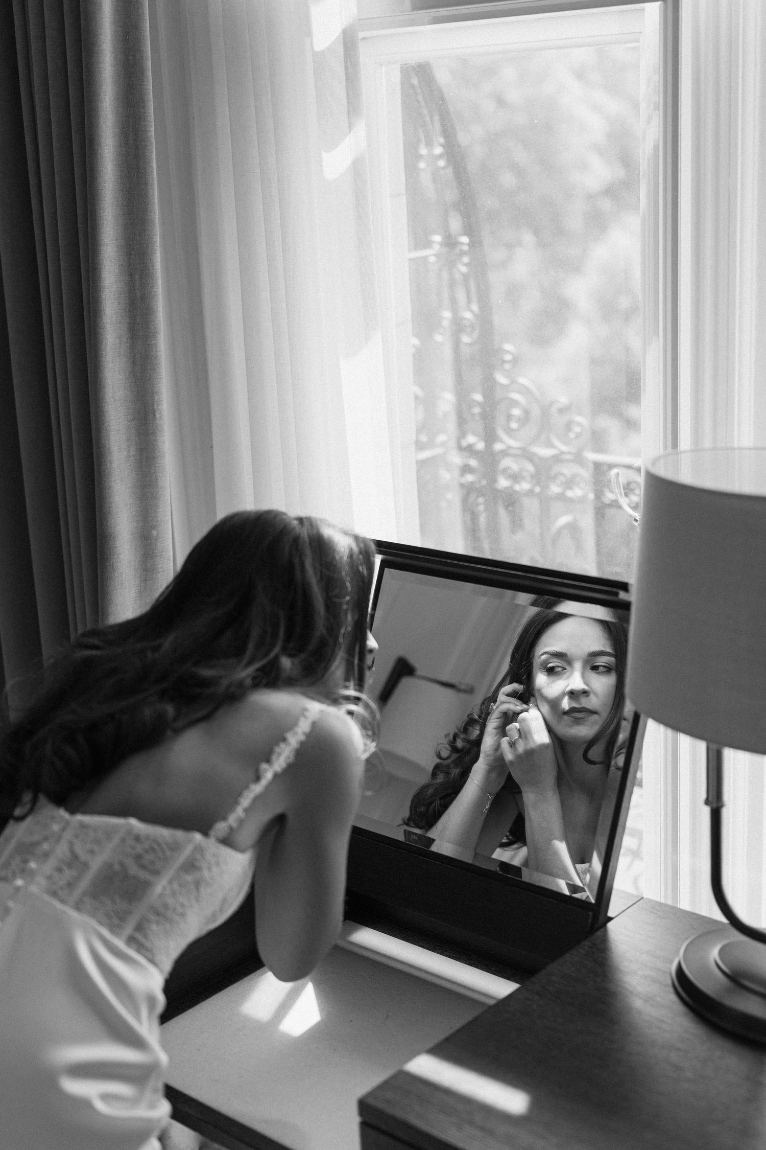 Black and white photo of a bride applying makeup while looking into a vanity mirror by a window.