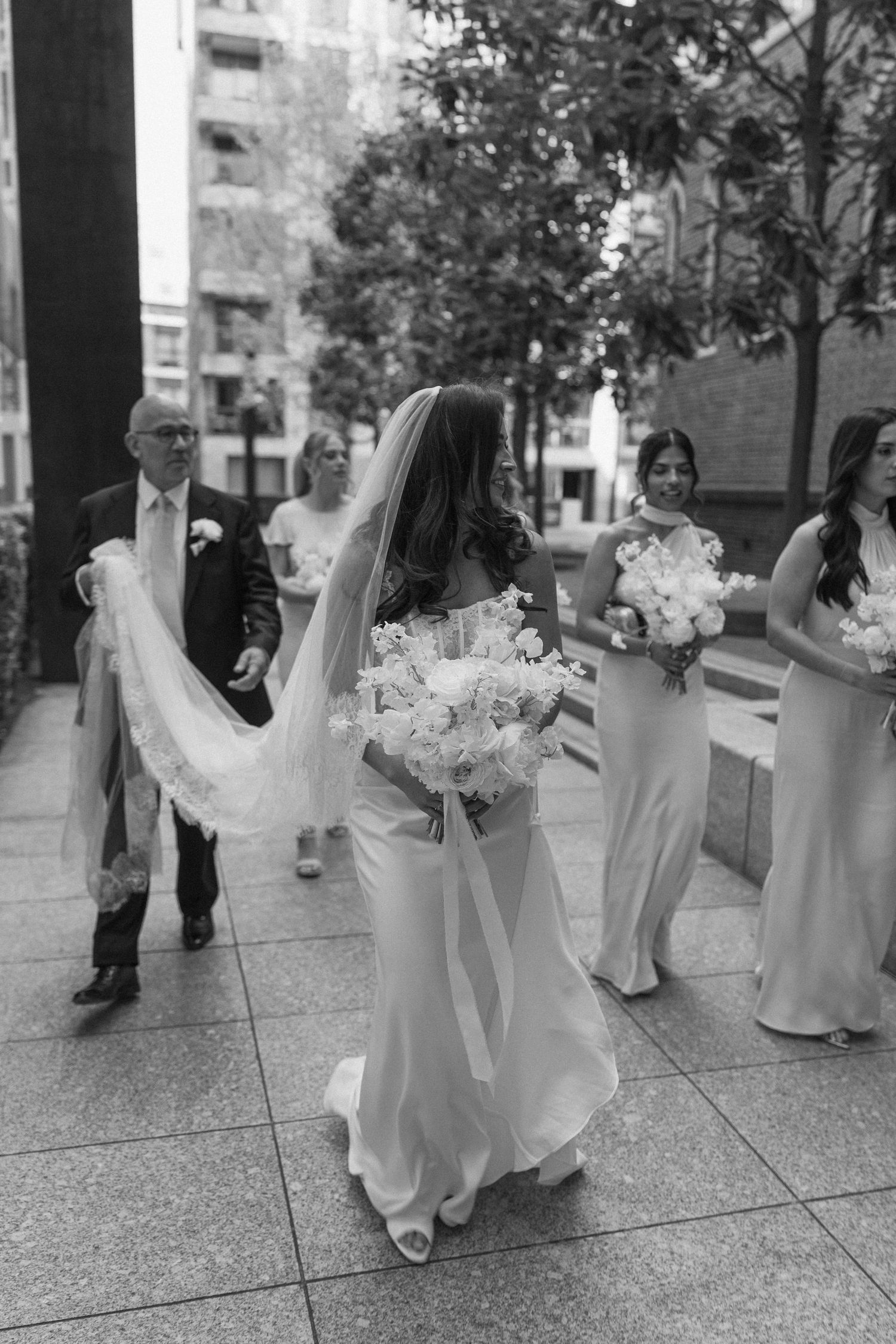 Black and white image of bride with bouquet and bridal party walking toward a church entrance.