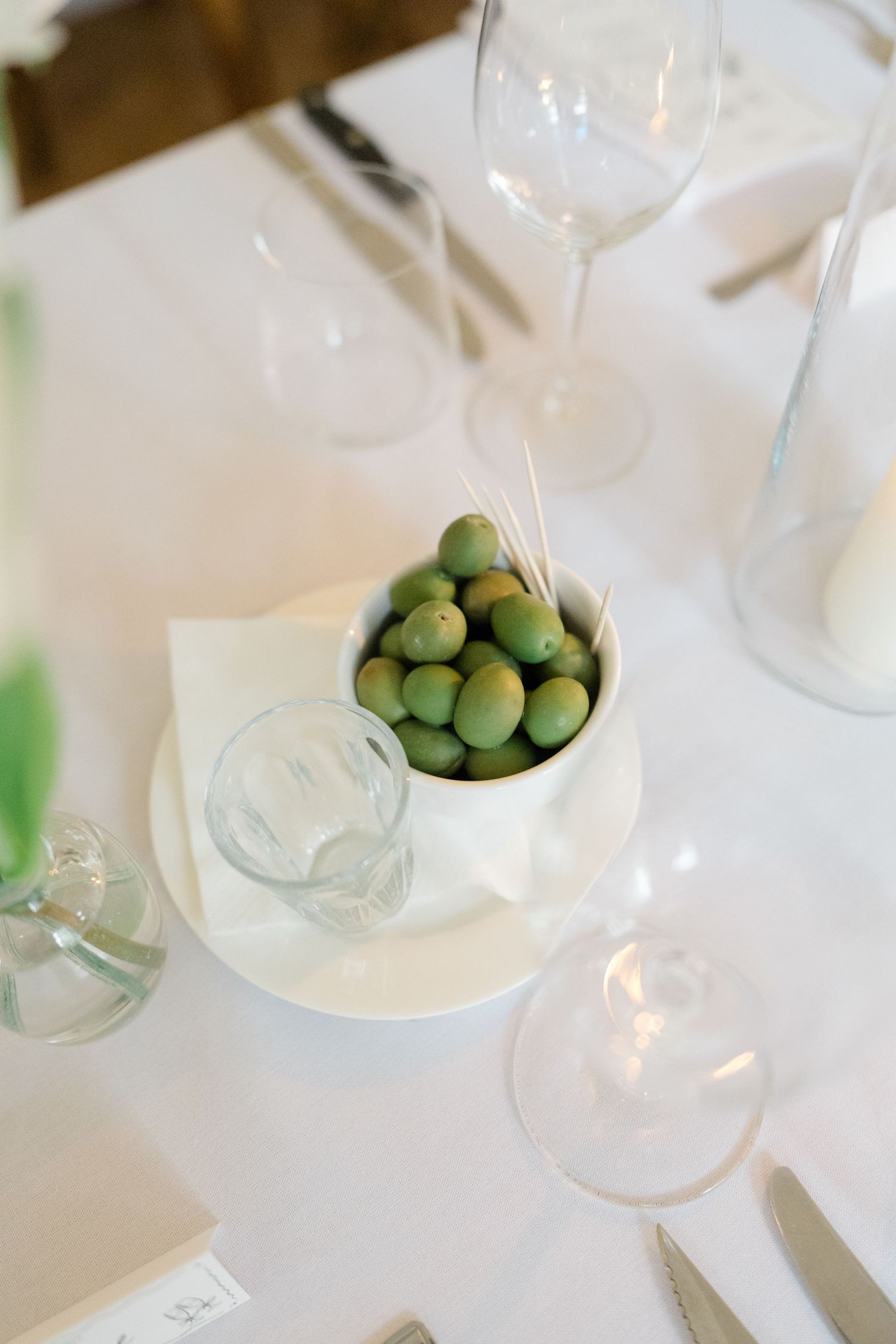 Elegant wedding reception table setting with a bowl of green olives, wine glasses, and white linen.
