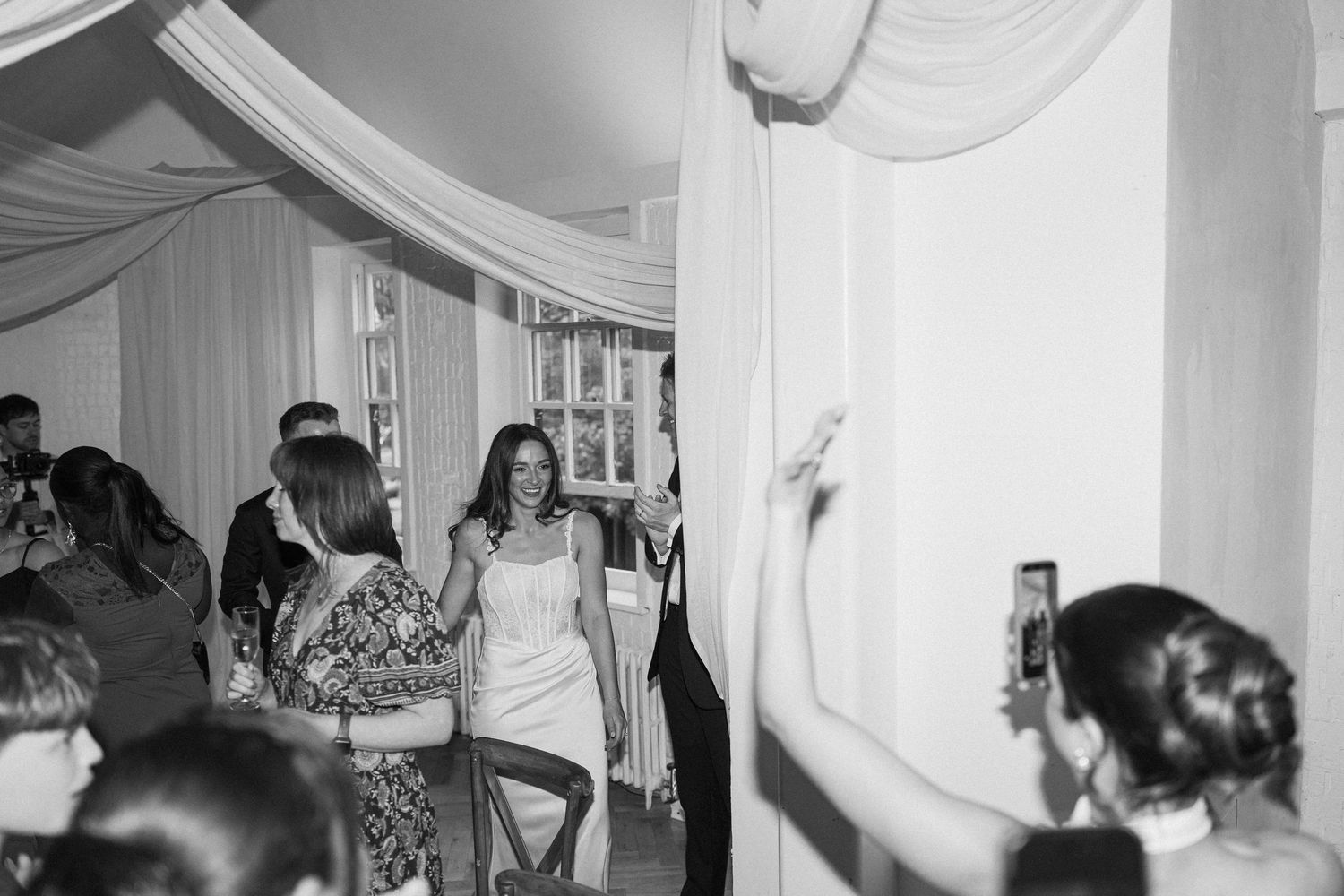 Black and white photo of bride walking through applauding wedding guests at indoor reception.