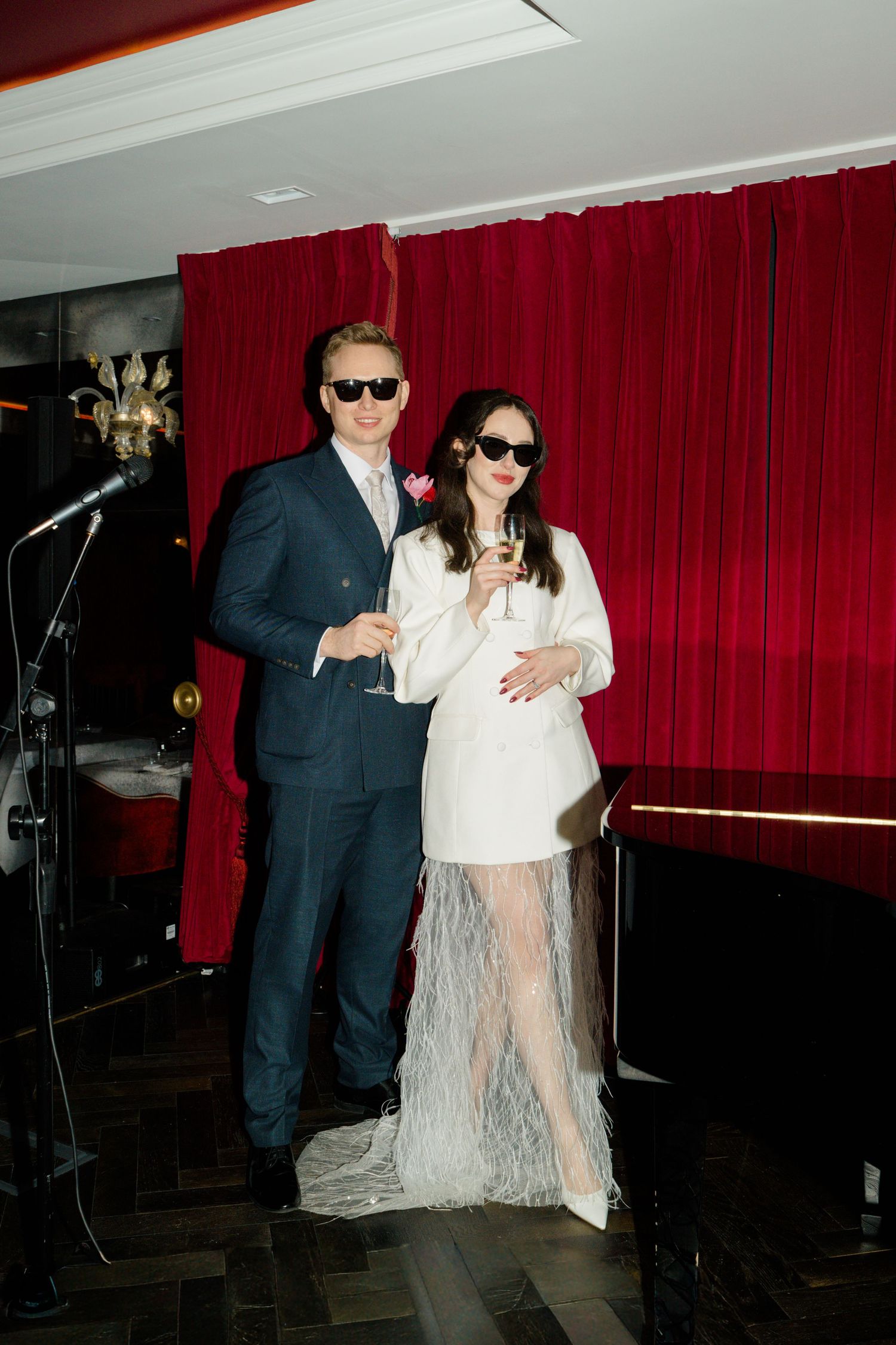 Newlyweds in sunglasses toast with champagne, bride in white blazer feather dress, groom in navy suit, red curtain backdrop.