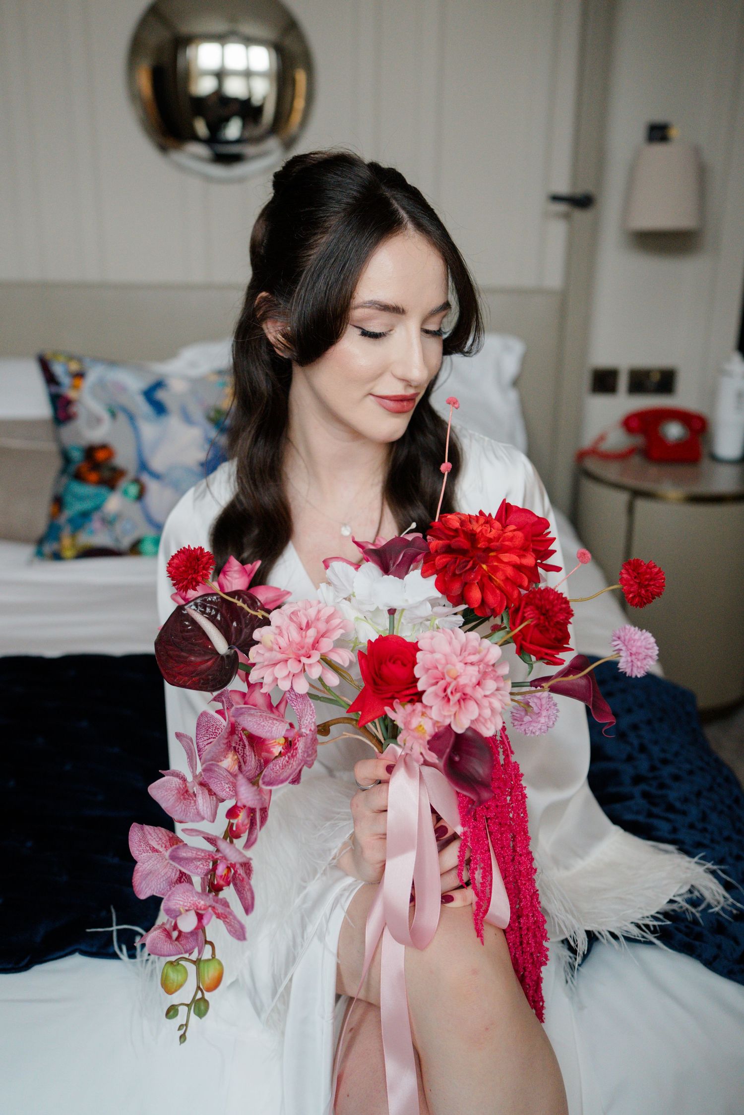 A dark-haired bride in a white robe sits on a bed holding a bouquet of red and pink flowers.