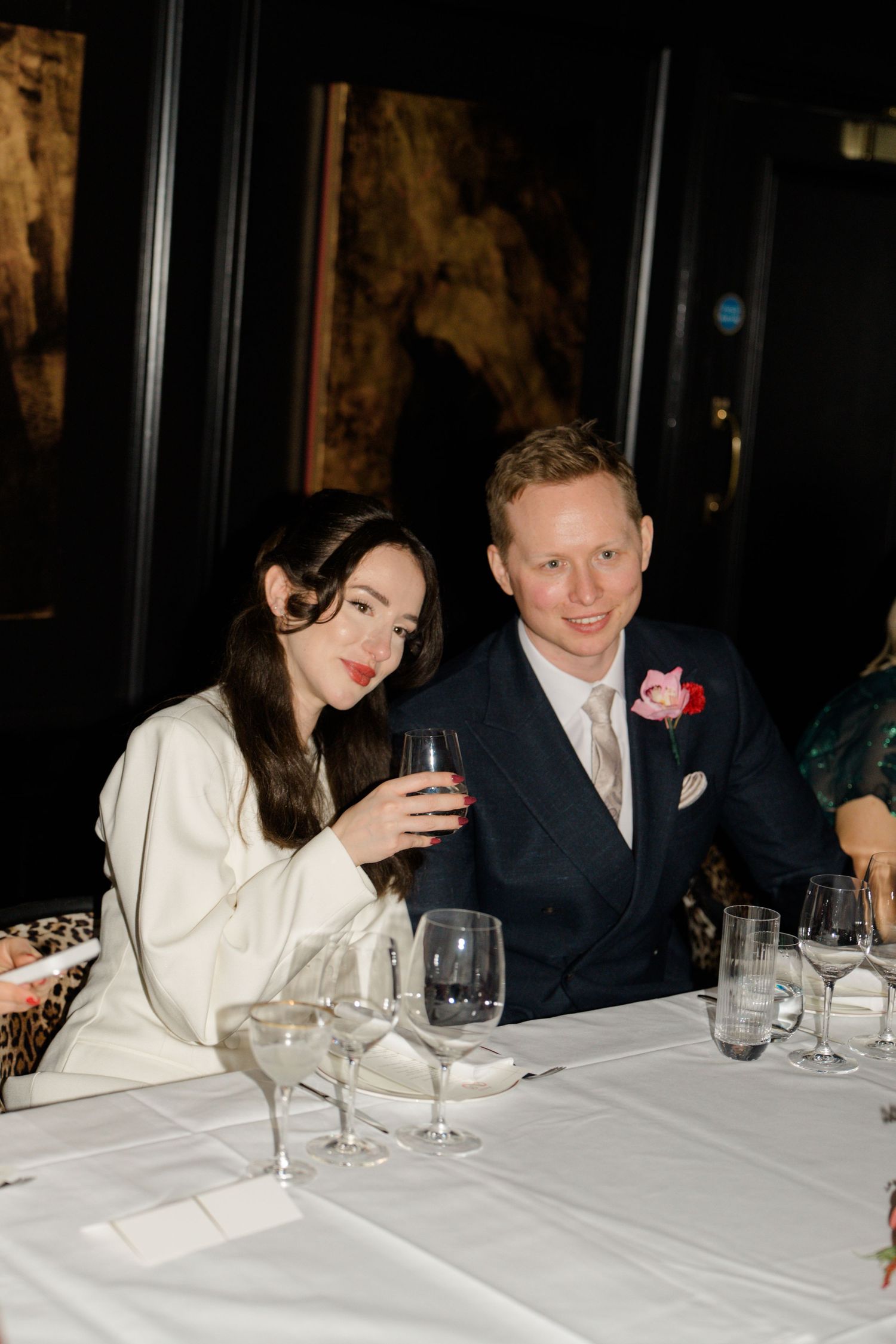 Bride in white blazer and groom in navy suit with pink boutonniere smile together at elegantly set wedding dinner table.