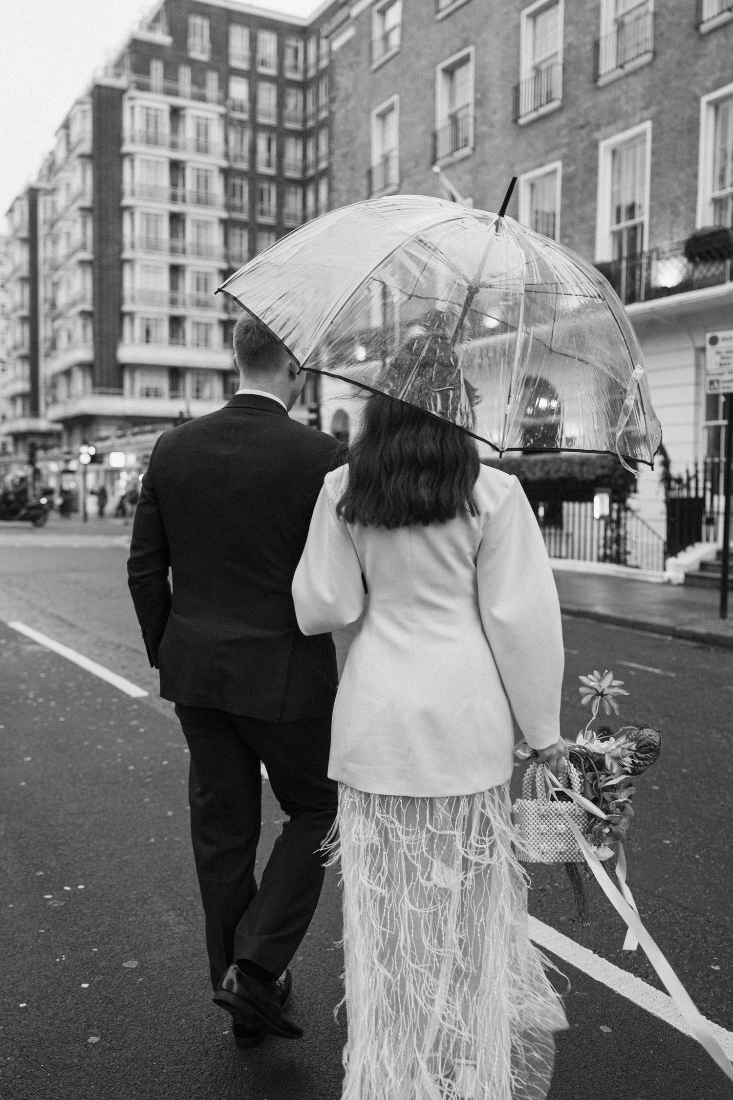 Bride and groom walking under clear umbrella on rainy London street, bride wearing feathered wedding dress.