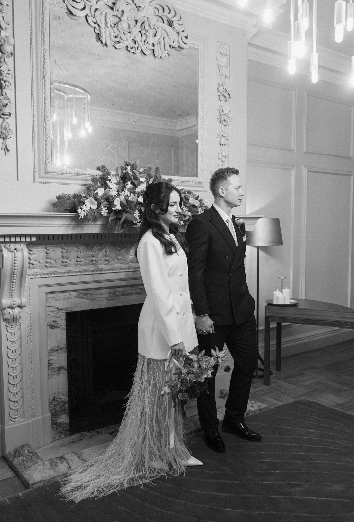 Bride and groom standing by ornate fireplace in black and white wedding photo.