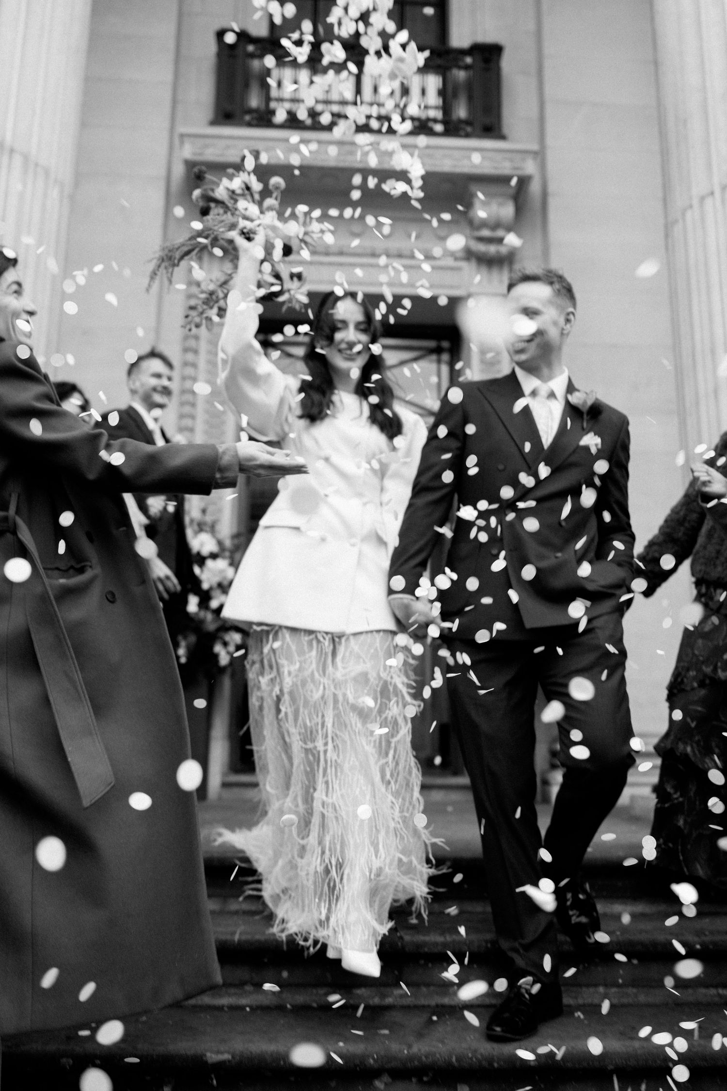 Black and white wedding confetti exit shot with bride in feather skirt and suited groom.