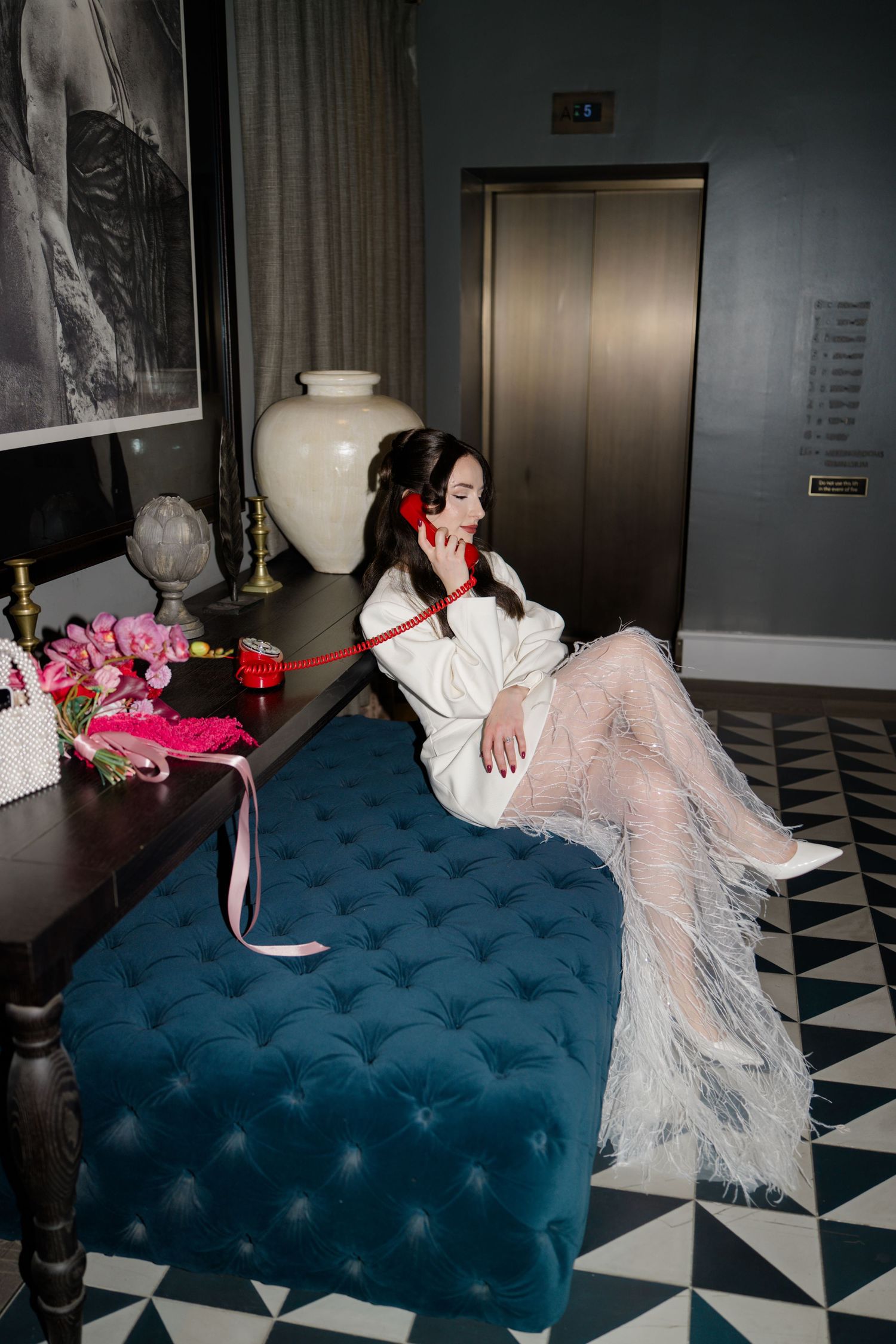 Elegant bride in white feather skirt talking on red telephone in luxury hotel suite.