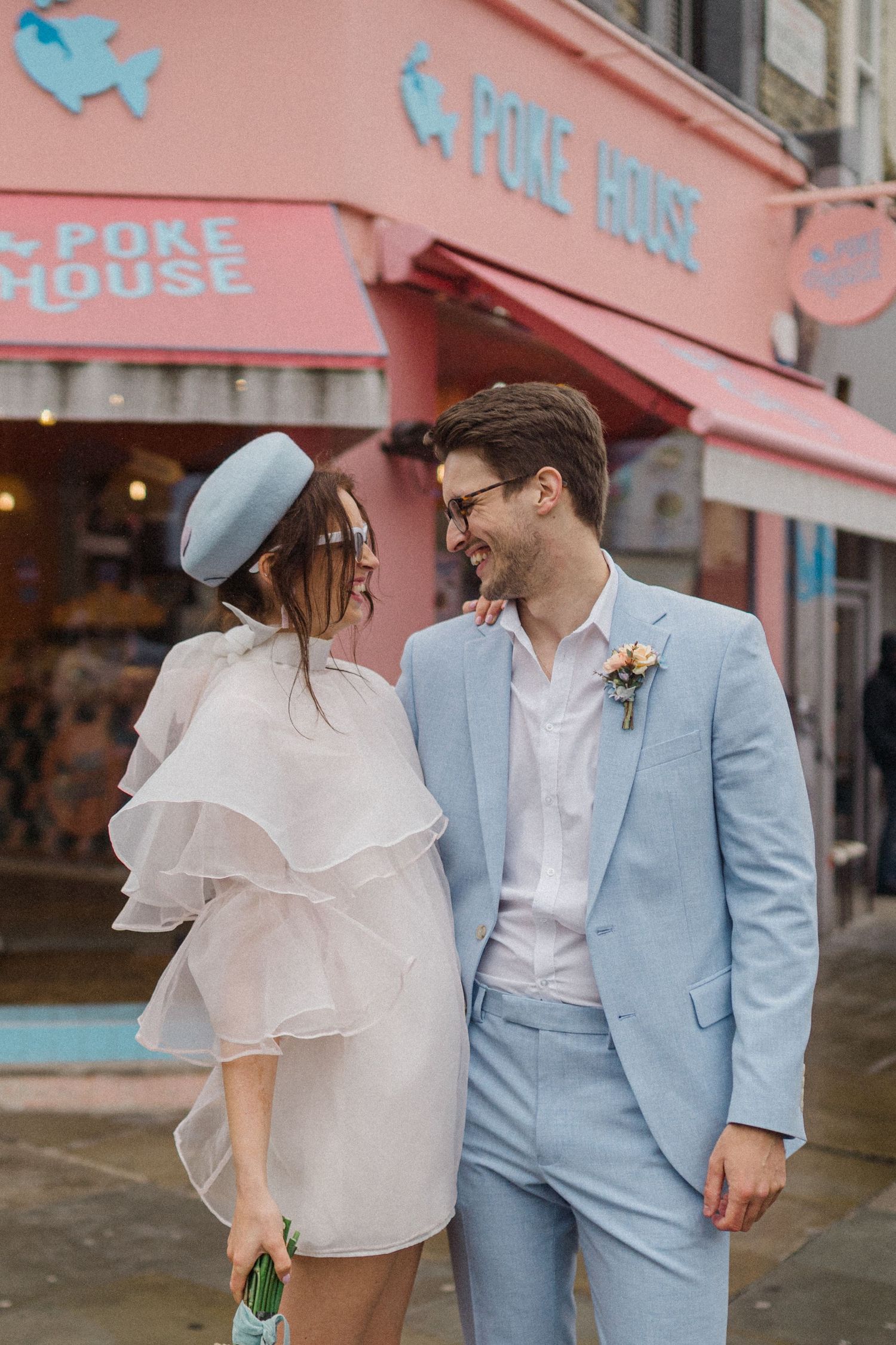 Couple in matching light blue outfits smiling outside a pink Poke House restaurant, bride in ruffled mini dress.