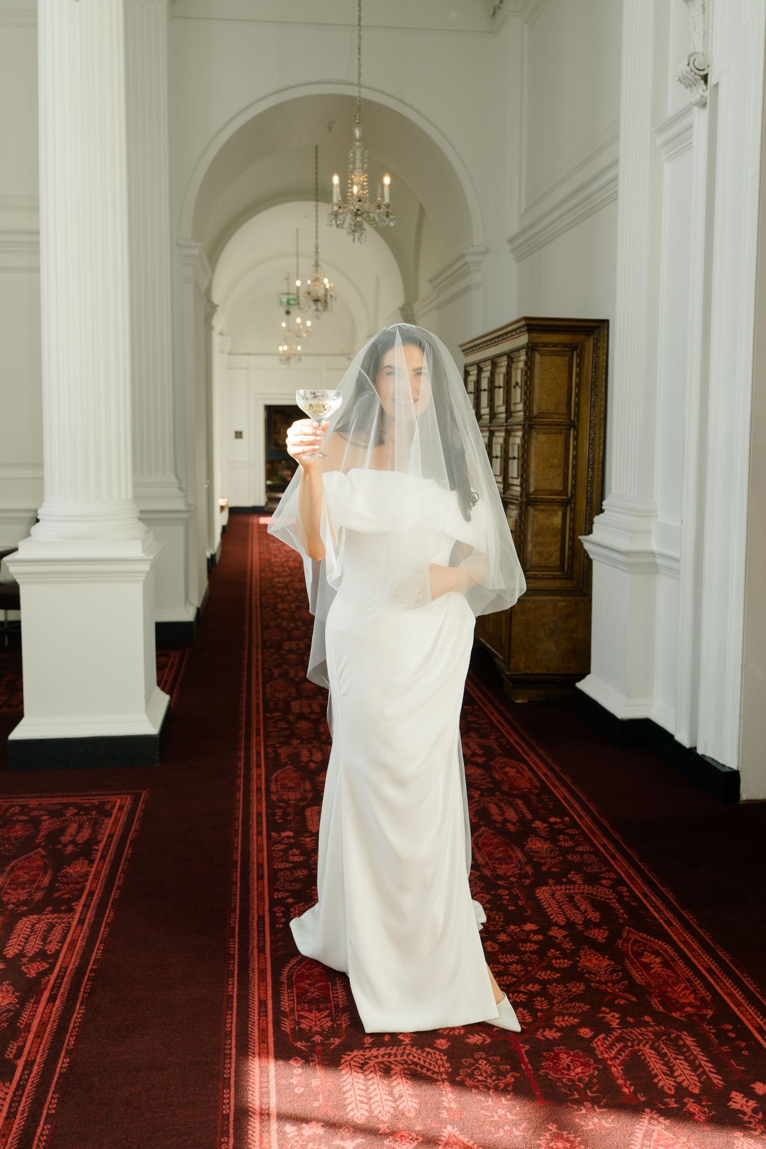 Bride in an off-shoulder white gown and sheer veil holds a champagne glass while walking down a grand hallway with a red patterned carpet.