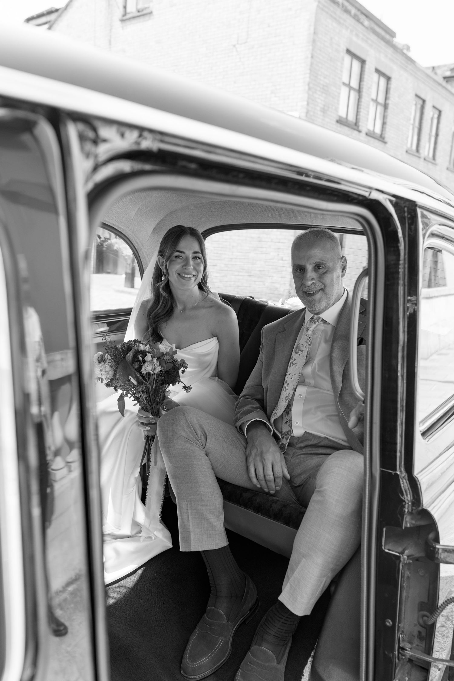 Black and white photo of smiling bride and father sitting together in vintage black taxi cab.