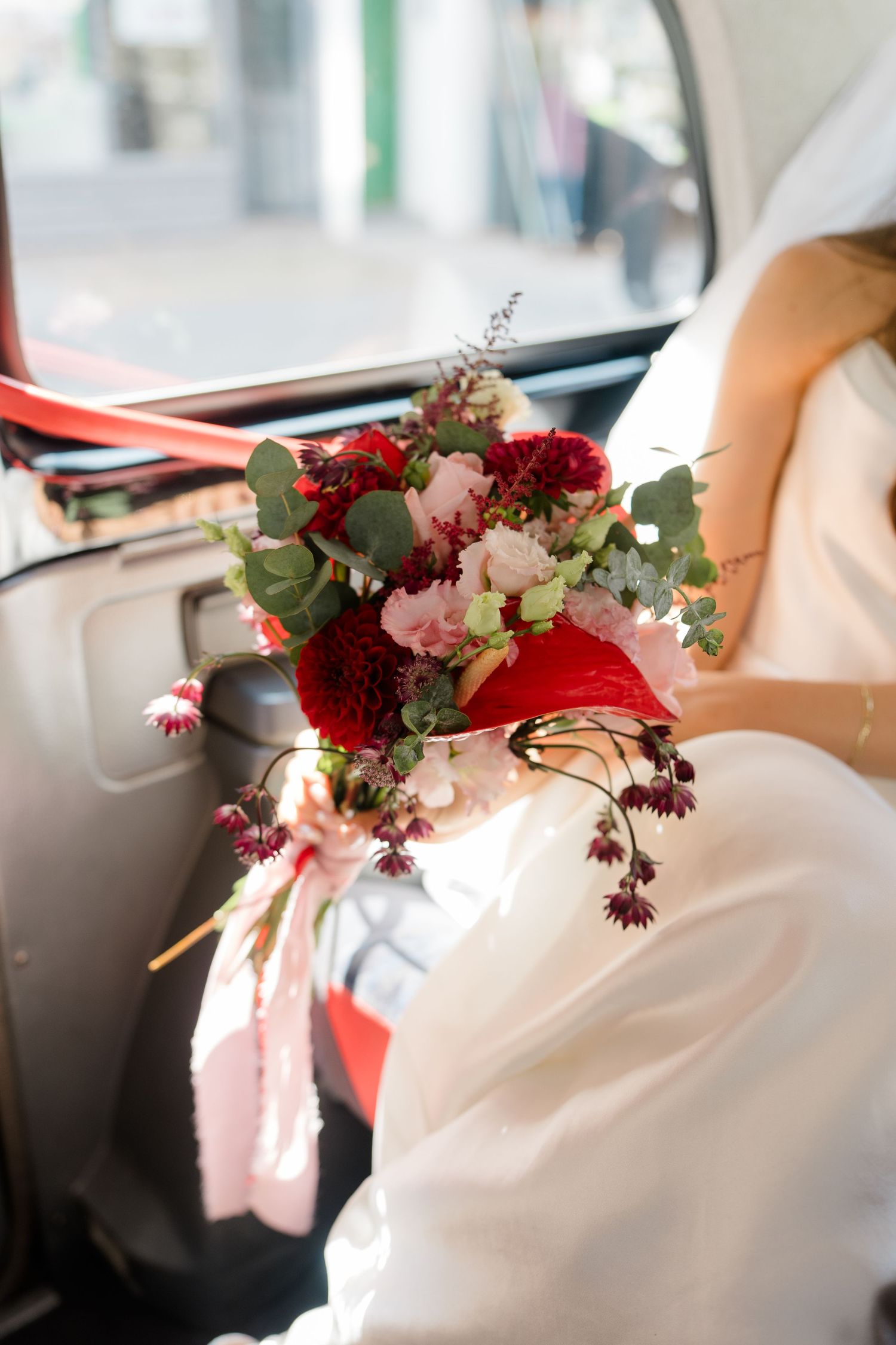 Bride in white dress holds vibrant red and pink floral bouquet while seated in a red taxi.
