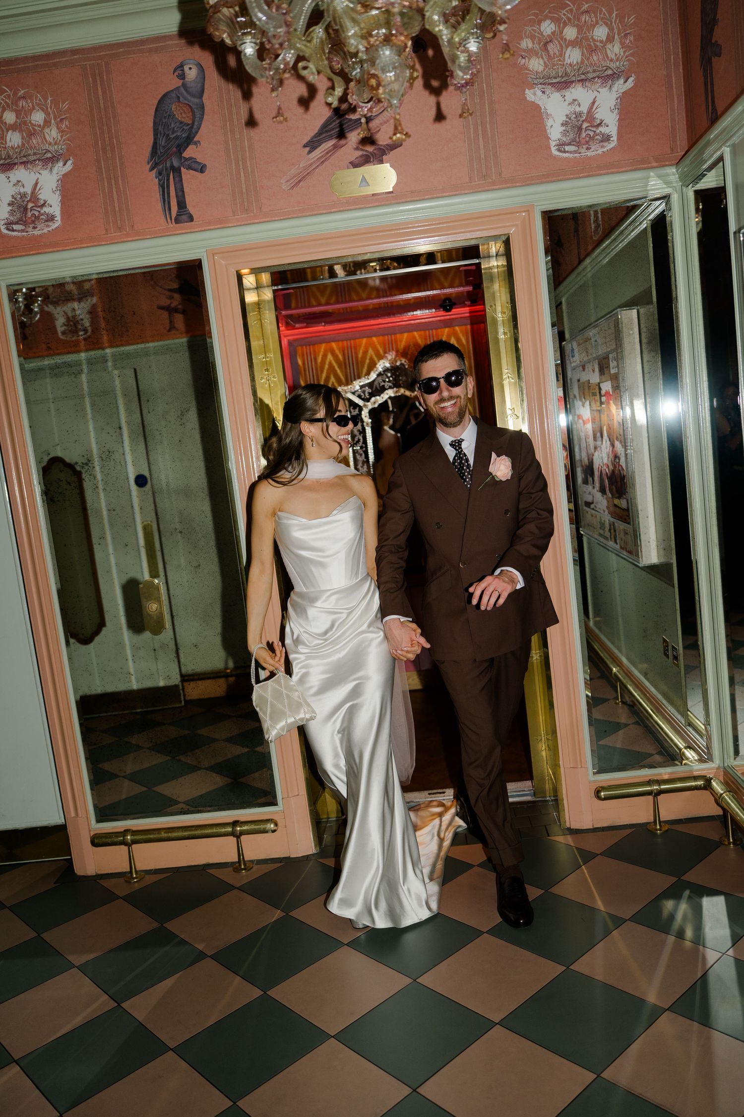 Bride and groom in sunglasses exit ornate elevator onto checkered floor in luxury hotel.