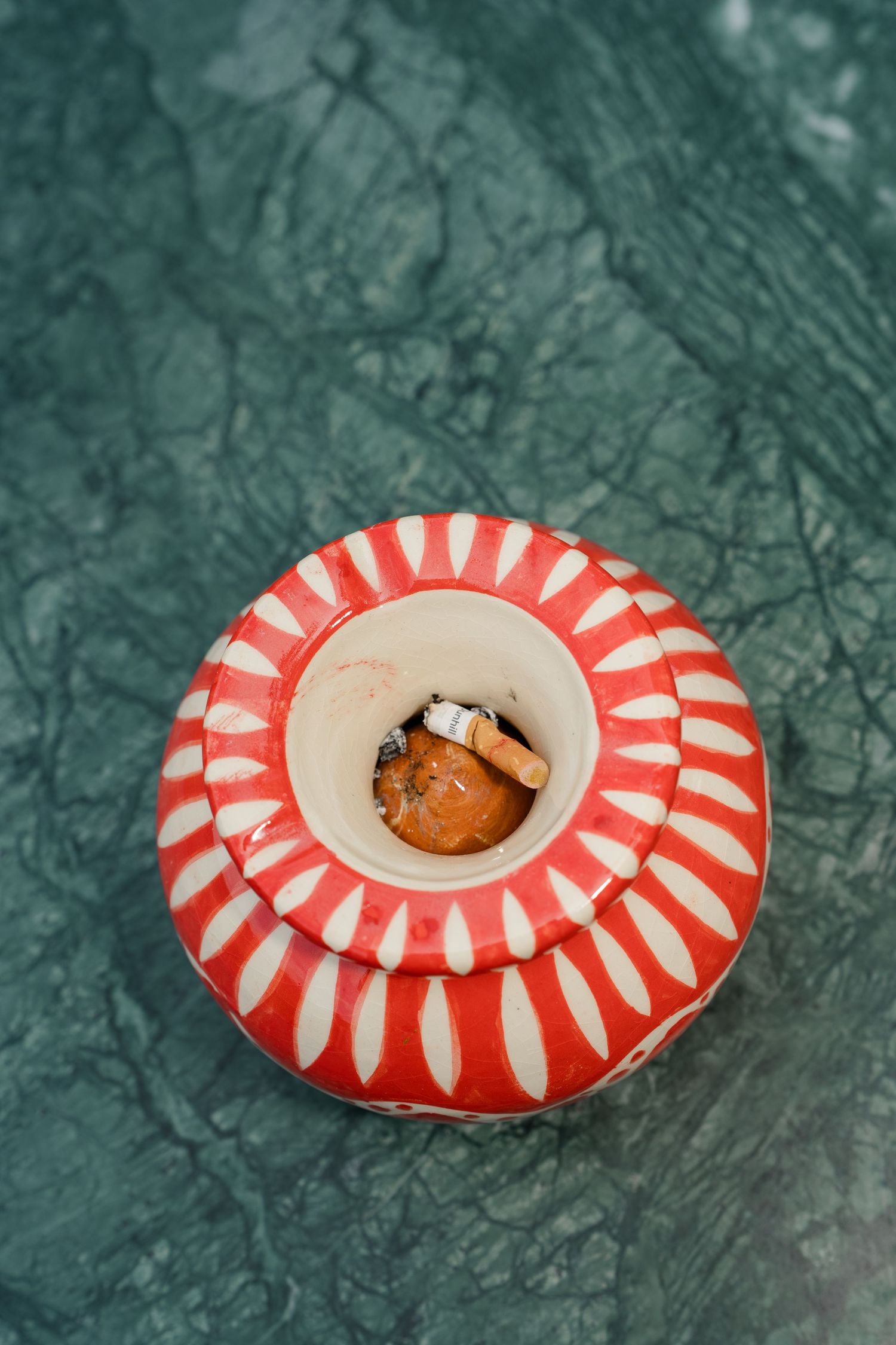 Red and white ceramic ashtray with cigarette butts on green marble surface.