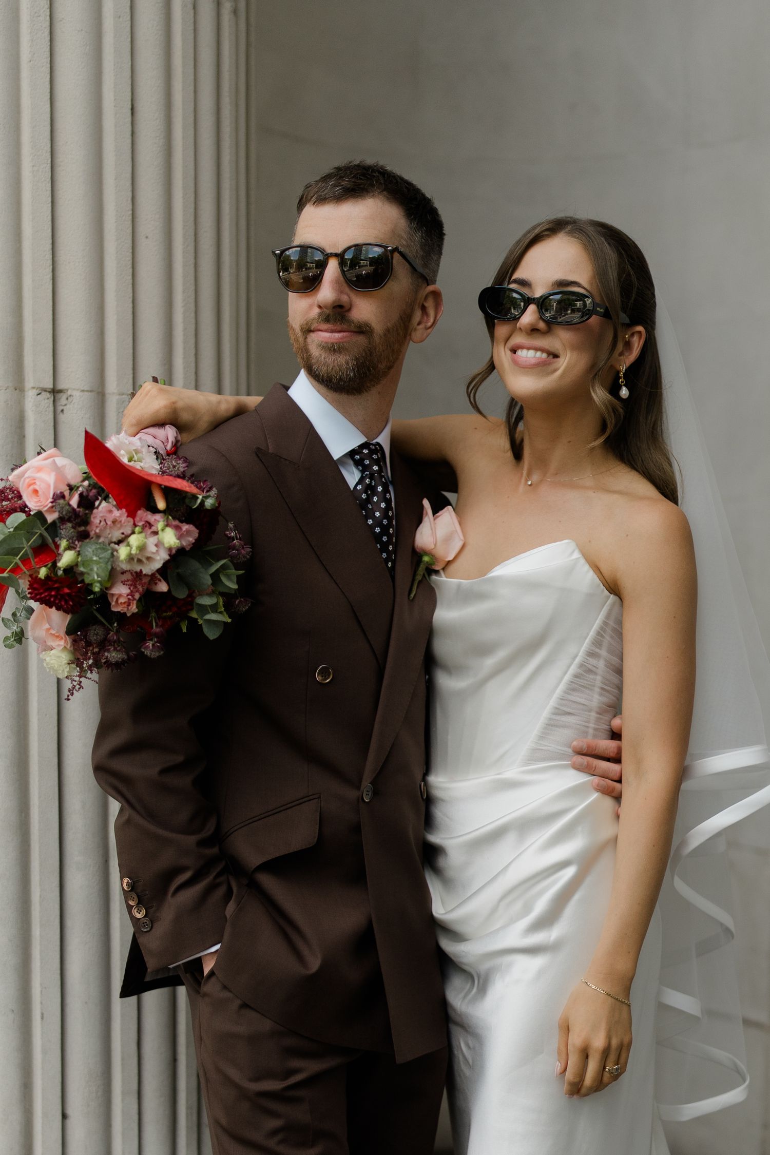 Stylish bride and groom pose in sunglasses outside a neoclassical building on their wedding day.