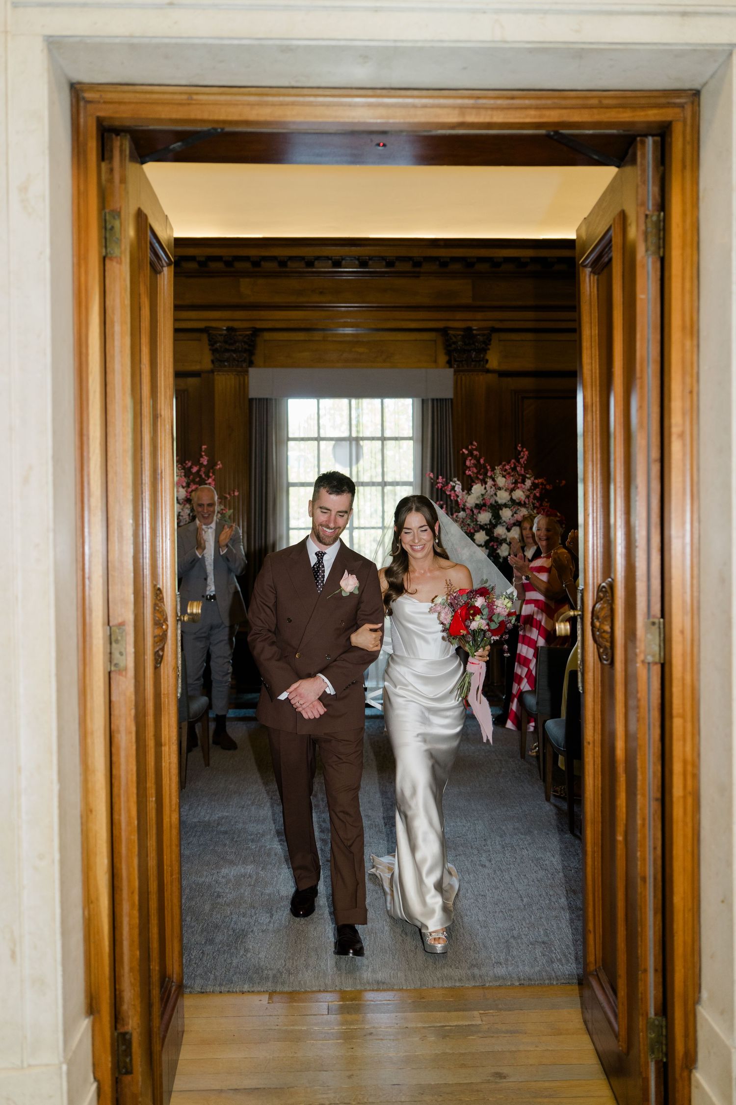 Bride and groom walk through wooden doors after wedding ceremony, bride in white satin gown holding red bouquet.