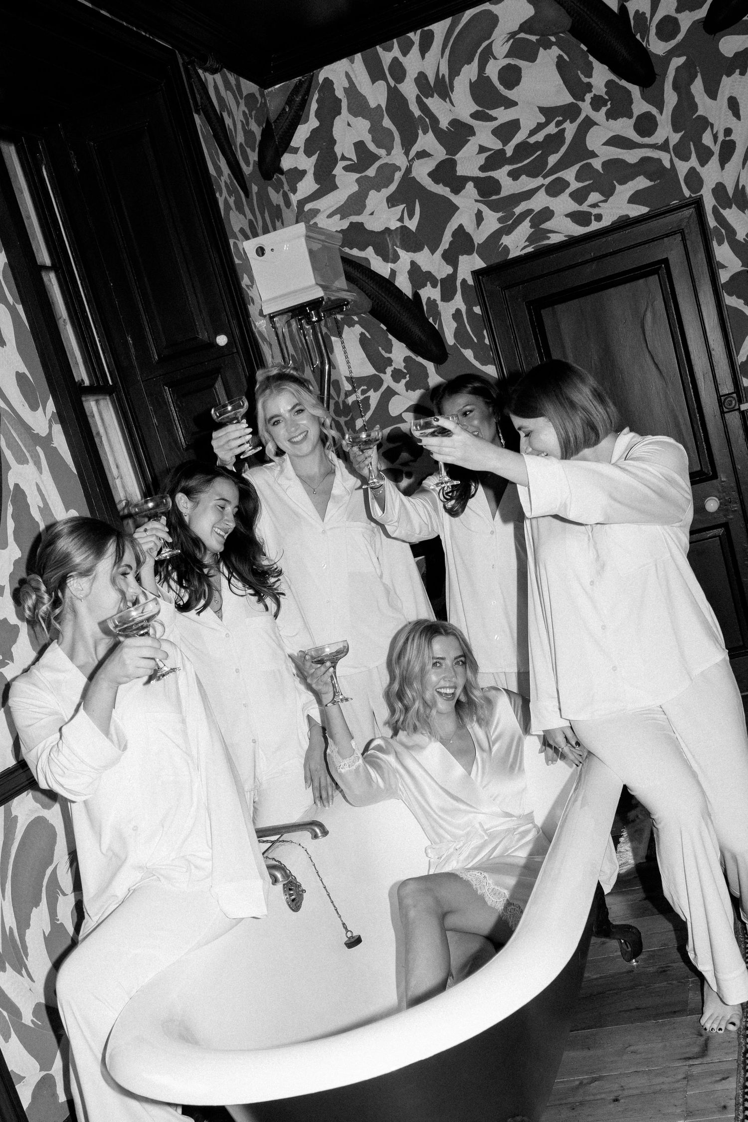Bridesmaids in white pajamas celebrate with champagne around a clawfoot bathtub in a decorated room.
