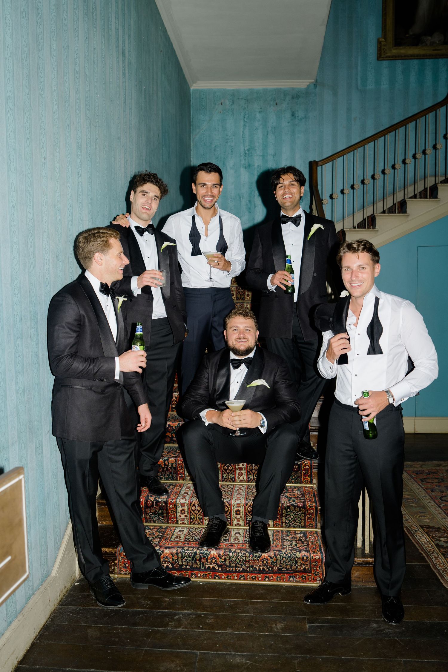 Groomsmen in black tuxedos pose on staircase holding drinks at wedding celebration.