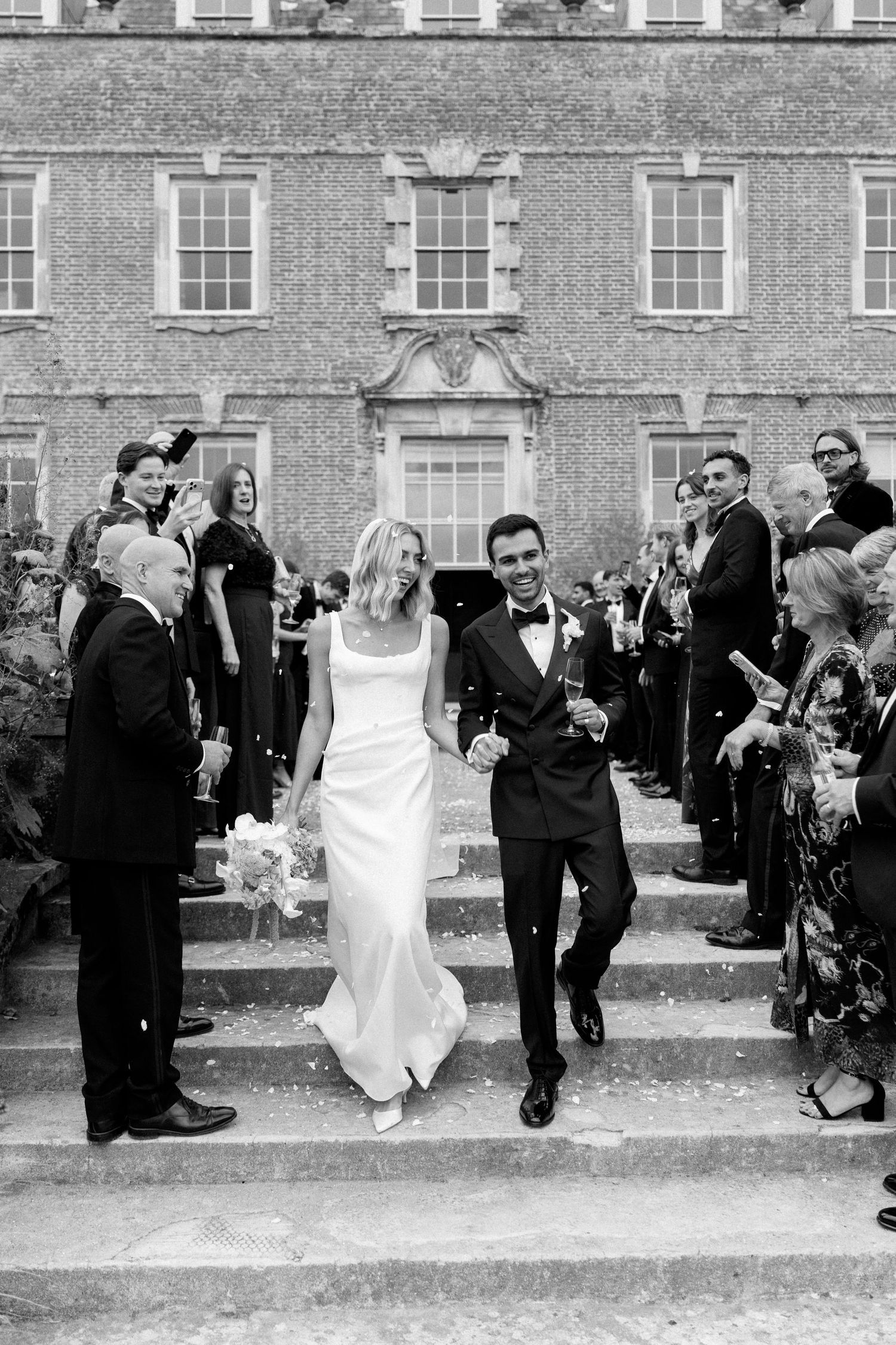 Bride and groom descend steps of grand brick manor house amid confetti in black and white photo.