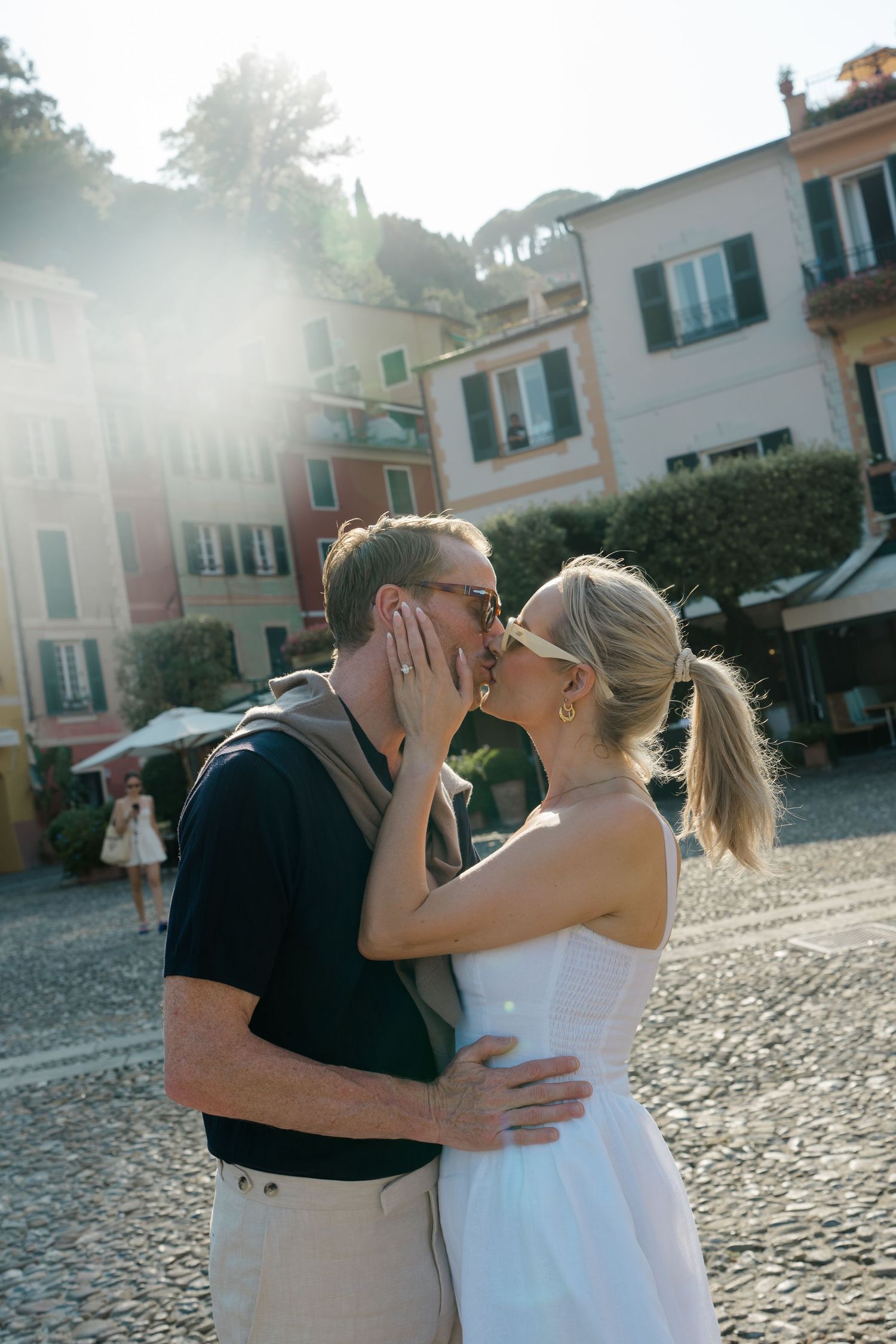 Couple sharing a romantic kiss in sun-drenched Portofino piazza, woman in white dress, colorful Italian buildings backdrop.