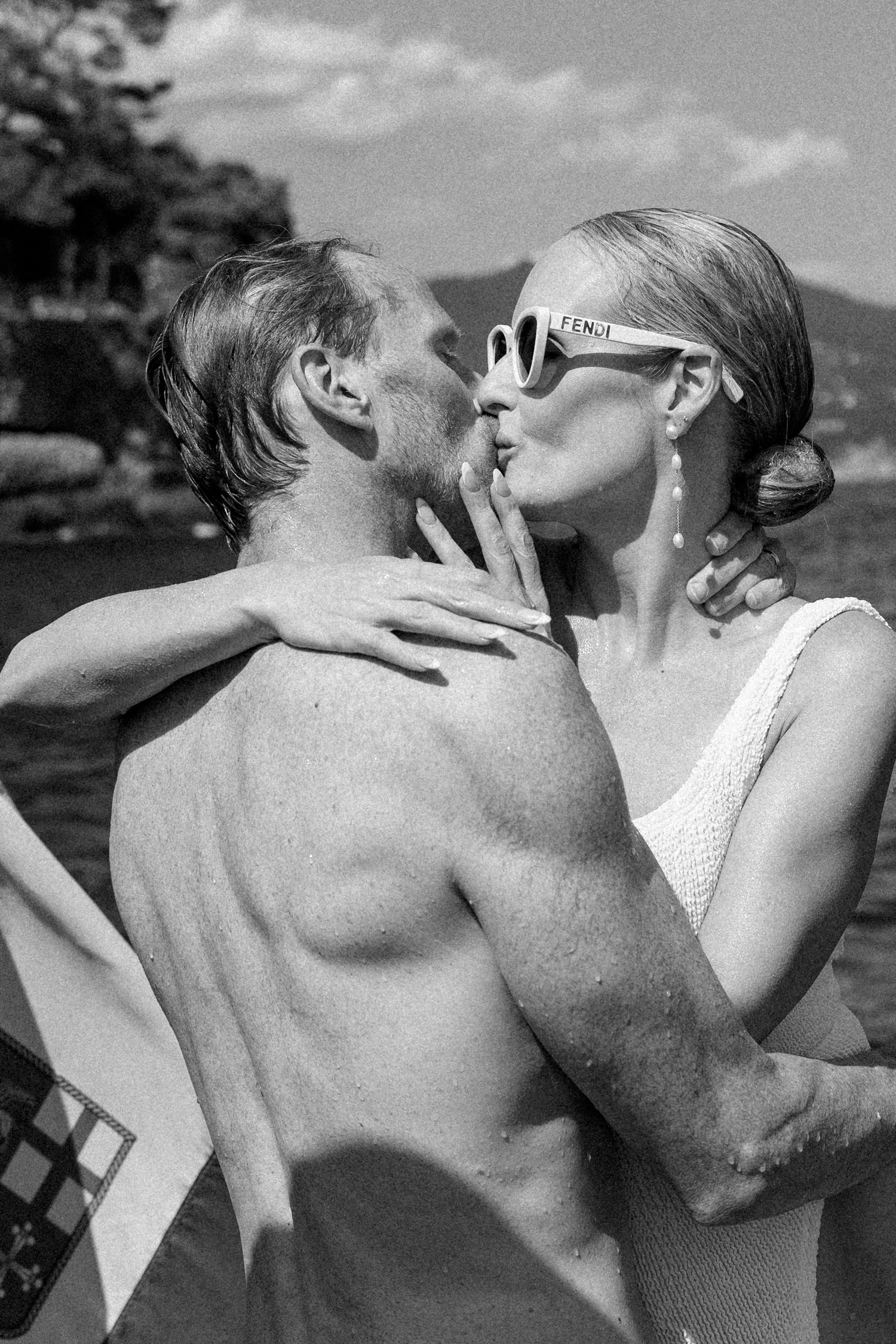 Black and white romantic photo of couple kissing wearing Fendi sunglasses near Italian lakeside after swimming.