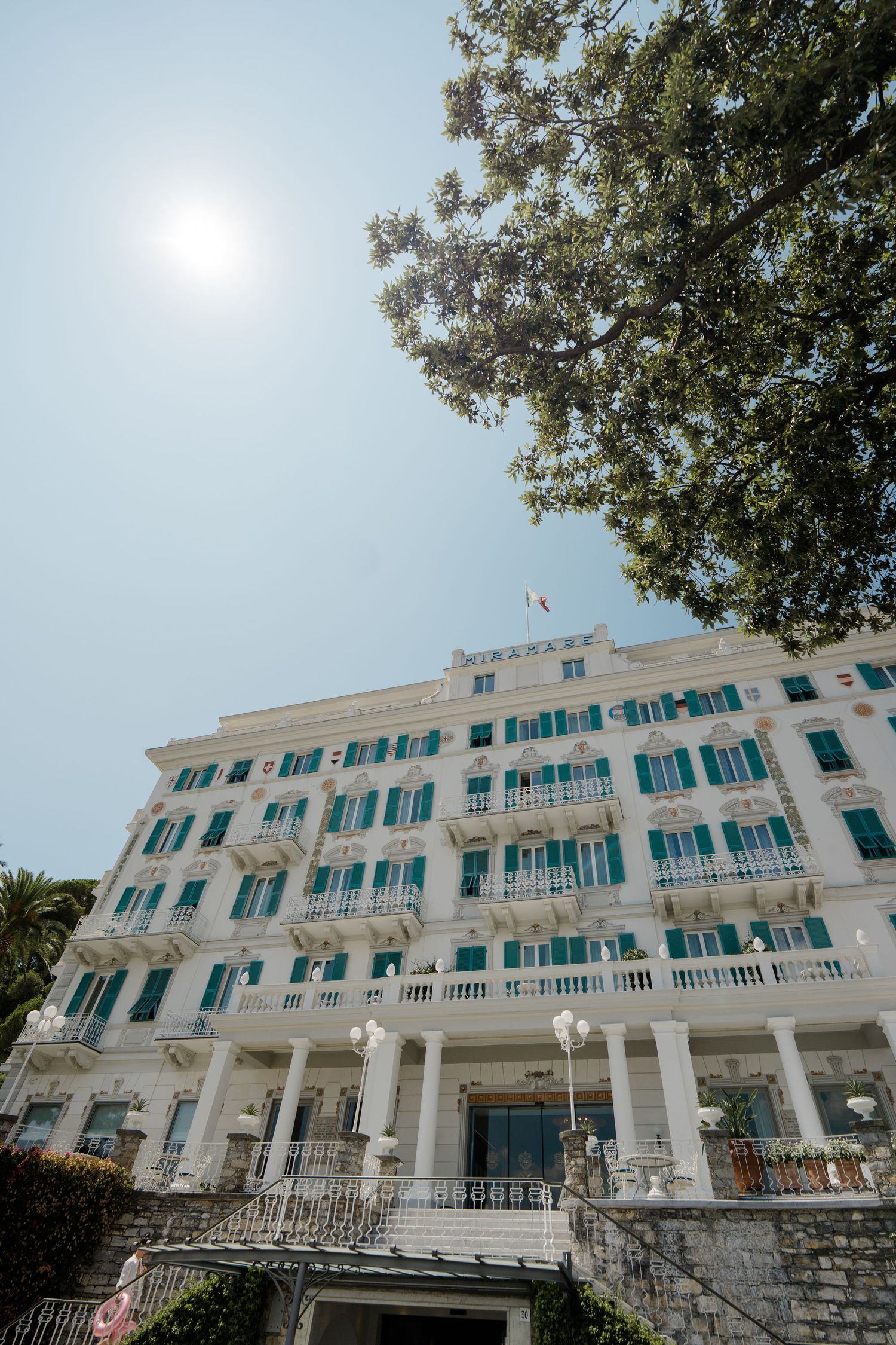 Grand white luxury hotel with green shutters and ornate balconies under bright sun in Italy.