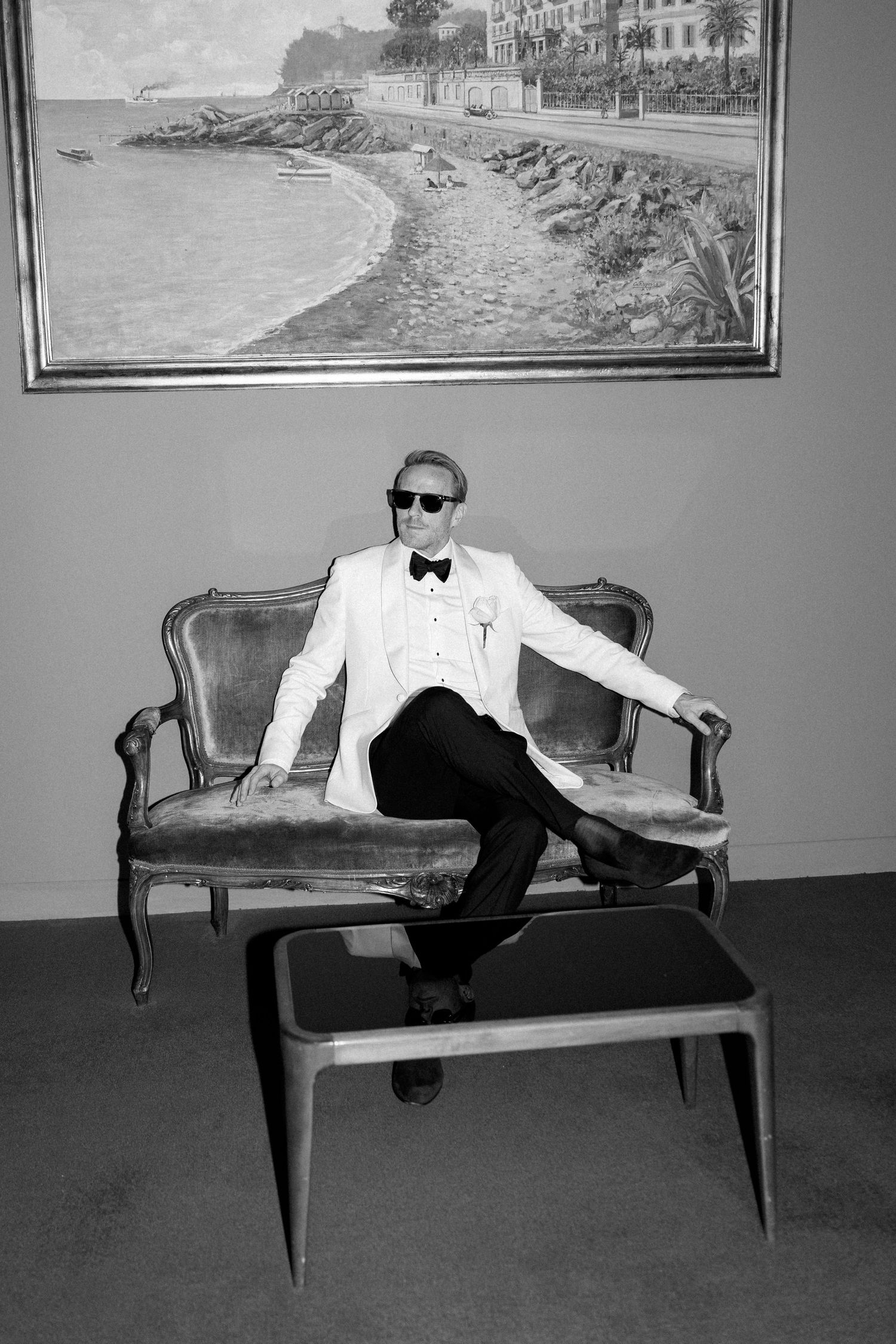 Stylish groom in white tuxedo and sunglasses posing on vintage sofa beneath coastal landscape painting.