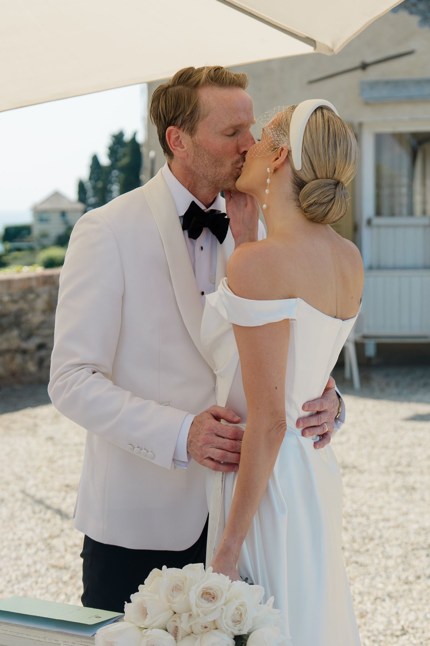Bride and groom share a kiss outdoors, groom in white tuxedo, bride in off-shoulder white gown.