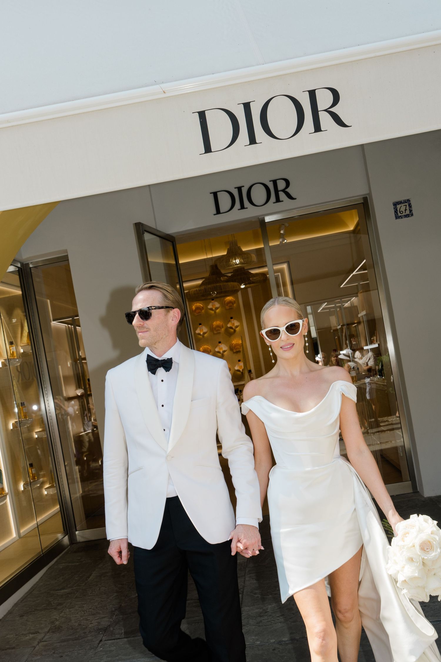 Elegant couple in white wedding attire holding hands walk out of Dior store wearing stylish sunglasses.