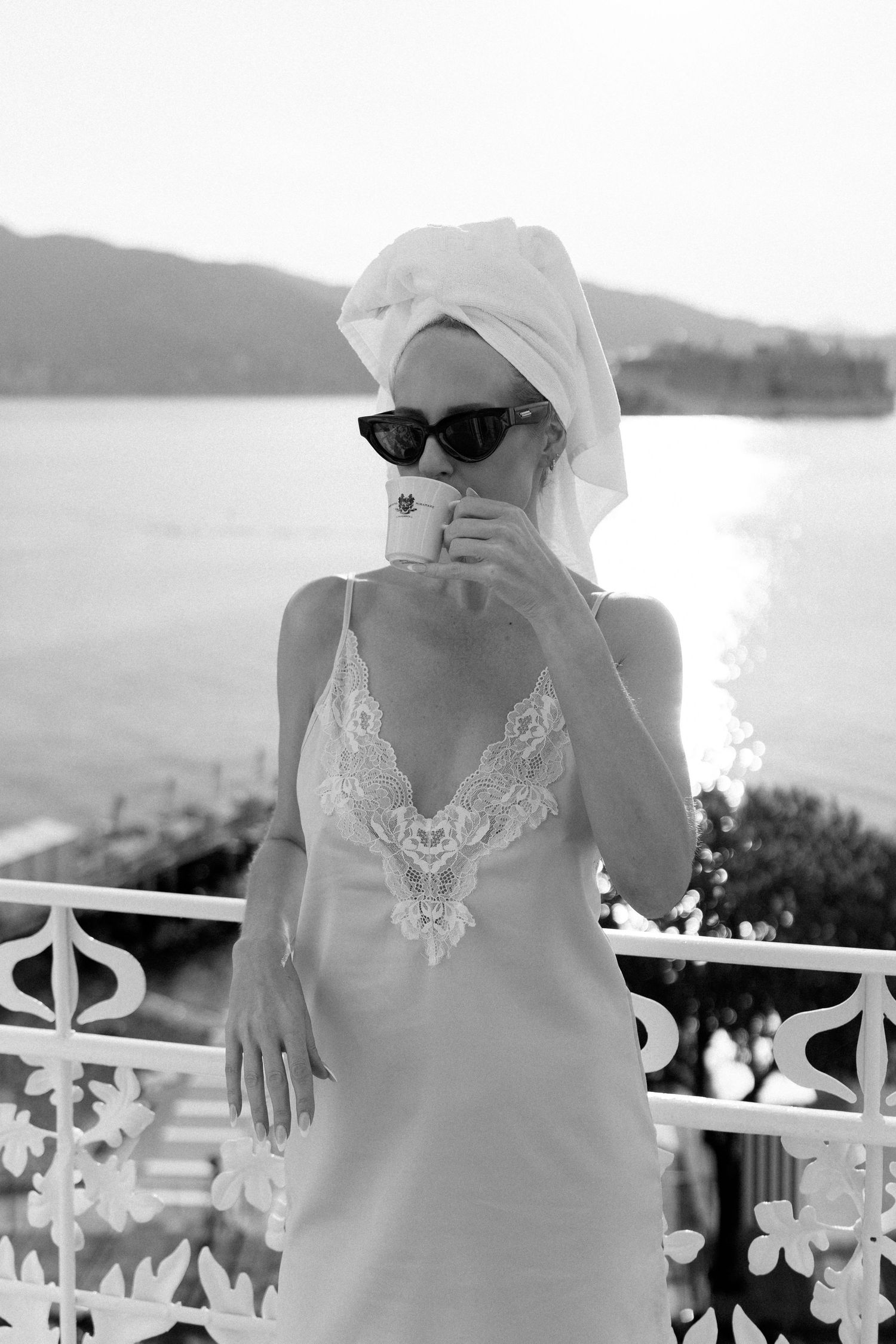 Black and white portrait of blonde woman in lace slip dress sipping coffee on balcony by sea.