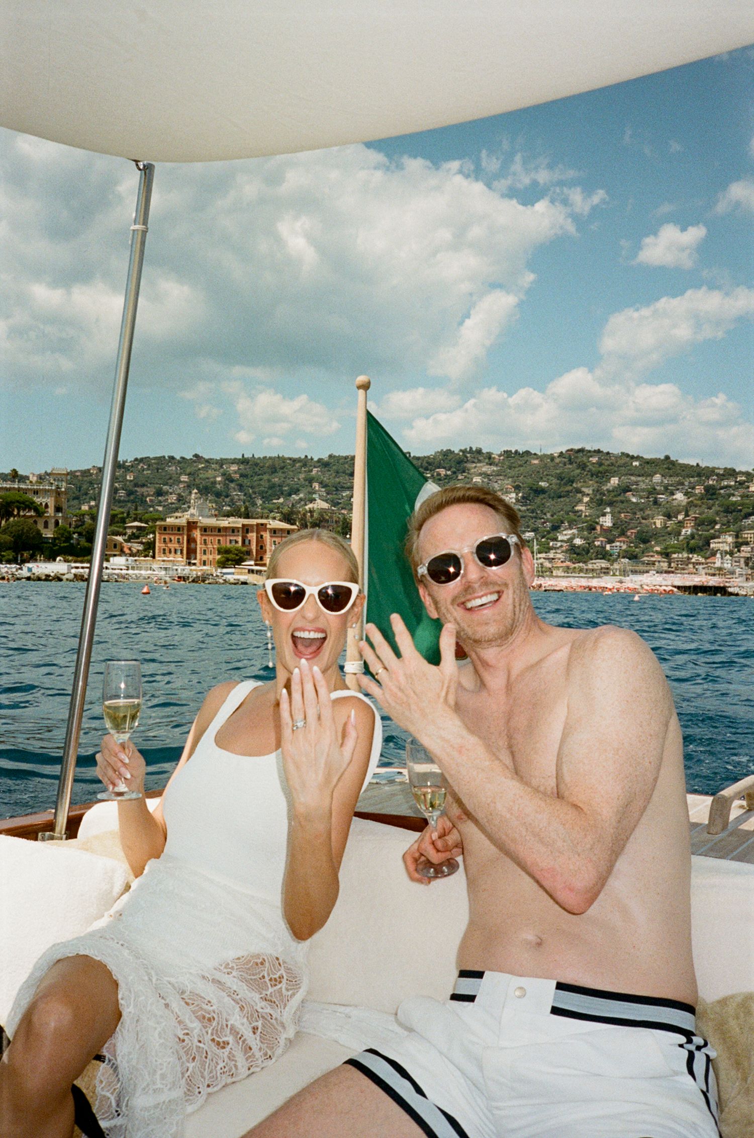 Engaged couple showing rings on luxury boat with champagne near French Riviera coastline.