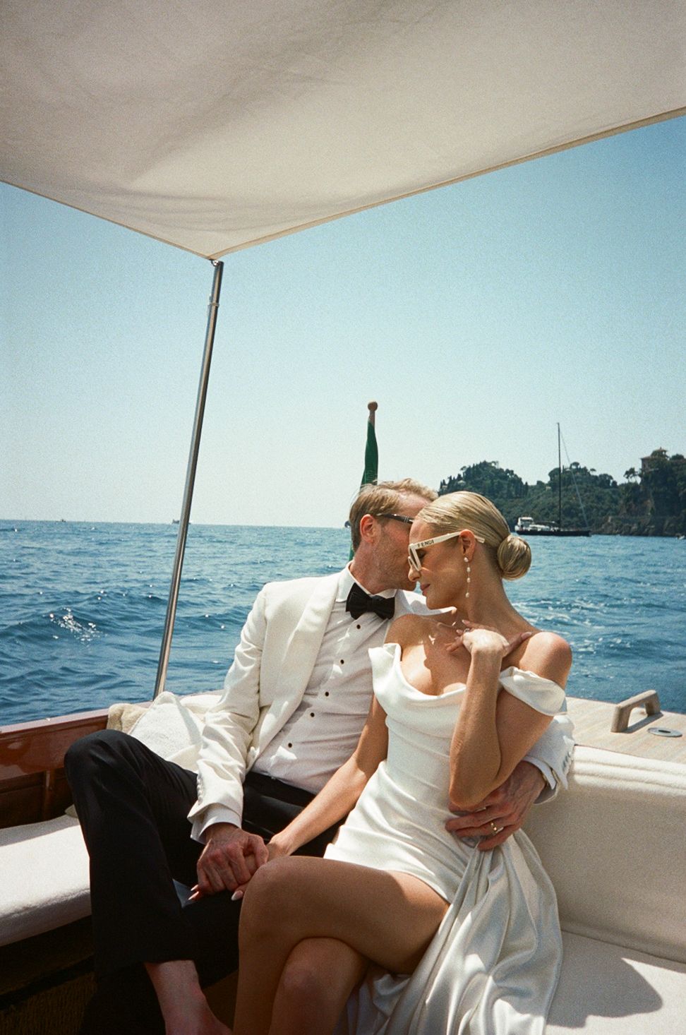 Bride and groom in wedding attire sitting closely together on a wooden boat sailing on blue Italian coastal waters.