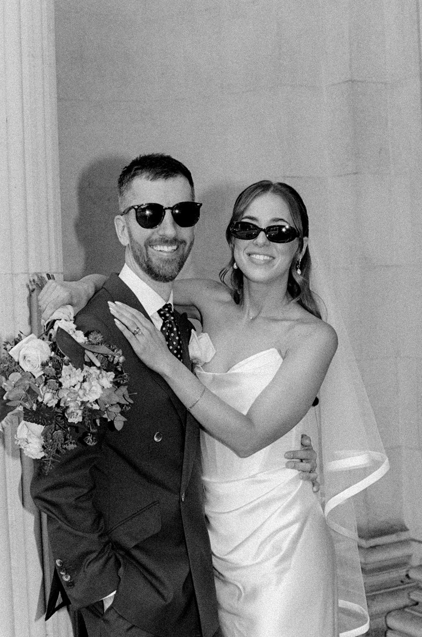 Smiling bride and groom in sunglasses pose together in black and white wedding portrait near stone columns.