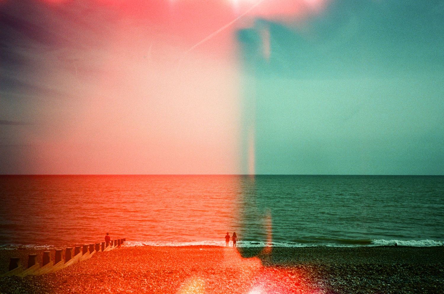 Split-toned beach photo with red and teal hues, silhouetted figures by the ocean under a dramatic sky.