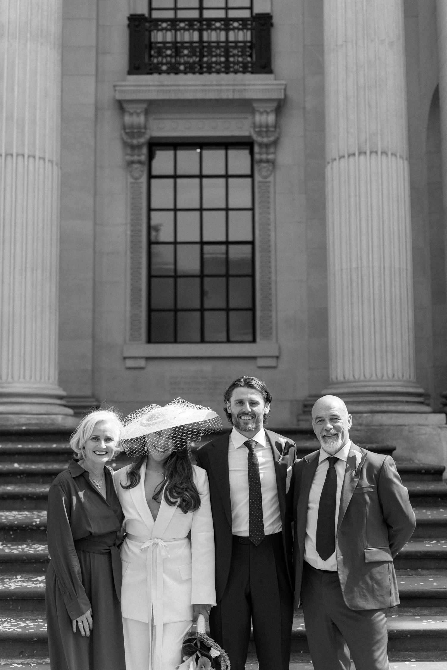 Black and white photo of smiling bride and groom with family outside grand columned building.