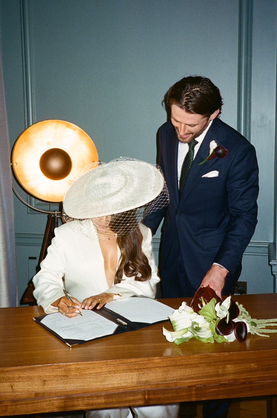 Bride in white suit and fascinator hat signs wedding documents as groom in navy suit looks on.