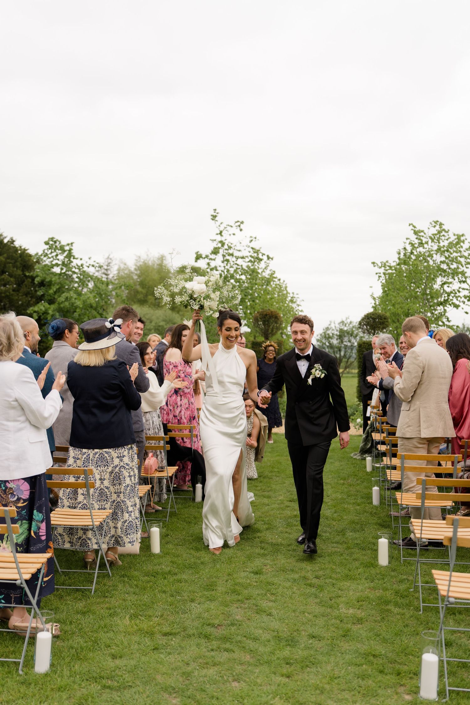 Bride and groom walk down outdoor aisle hand in hand as wedding guests celebrate with confetti.