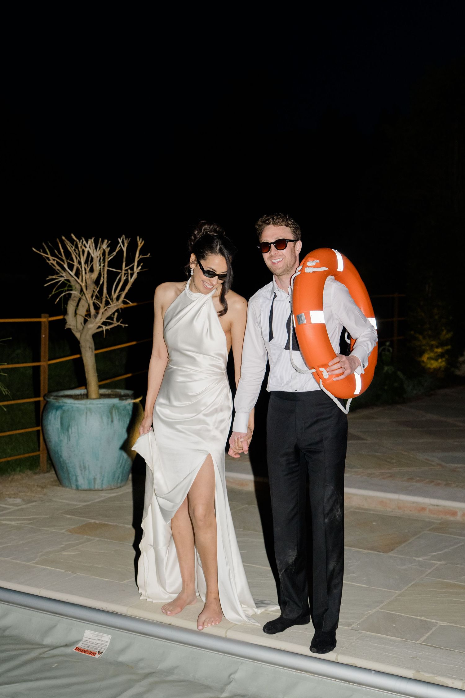Bride and groom in sunglasses stand poolside at night holding orange life preserver ring together.