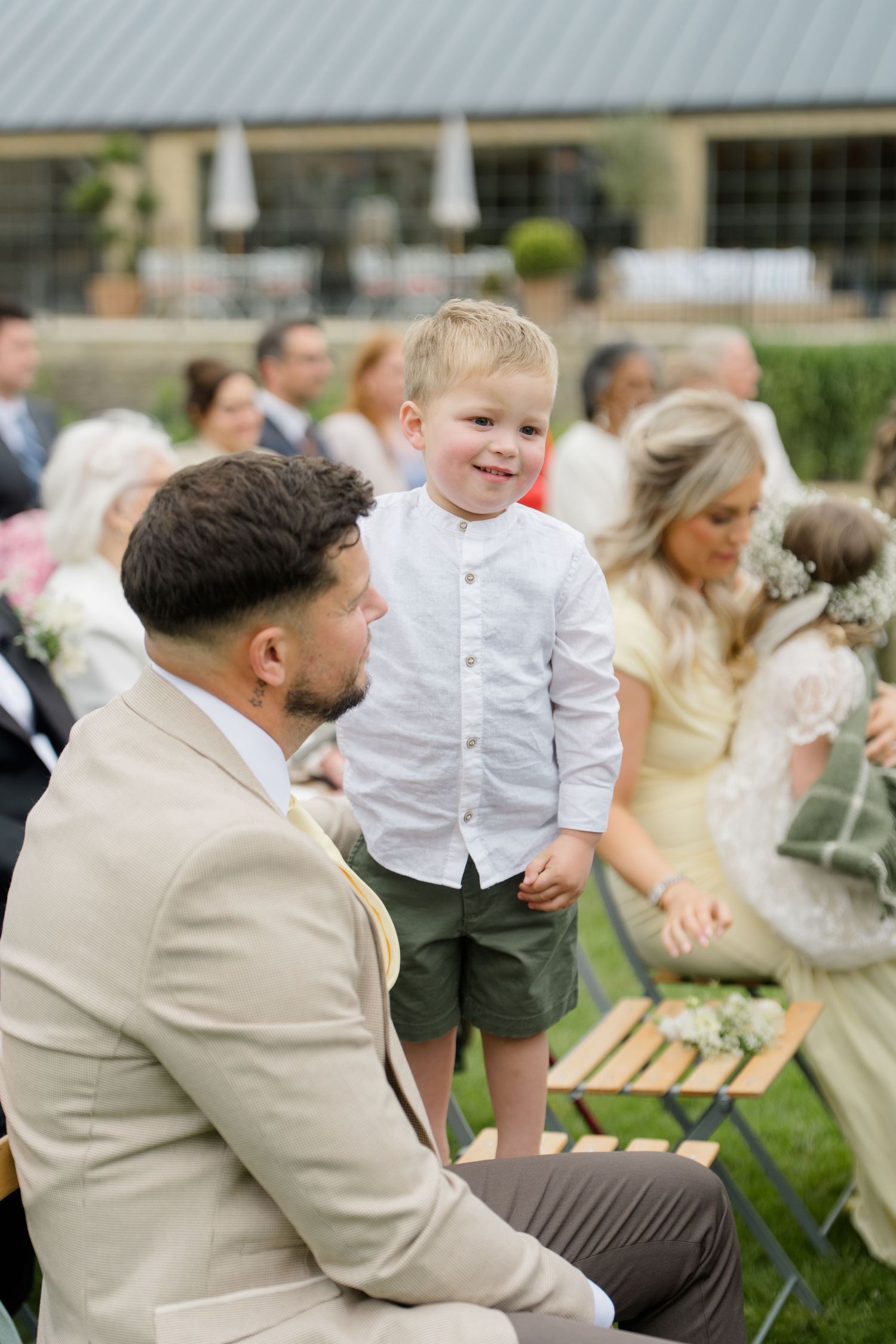 Smiling toddler boy in white shirt stands among seated wedding guests at outdoor ceremony.