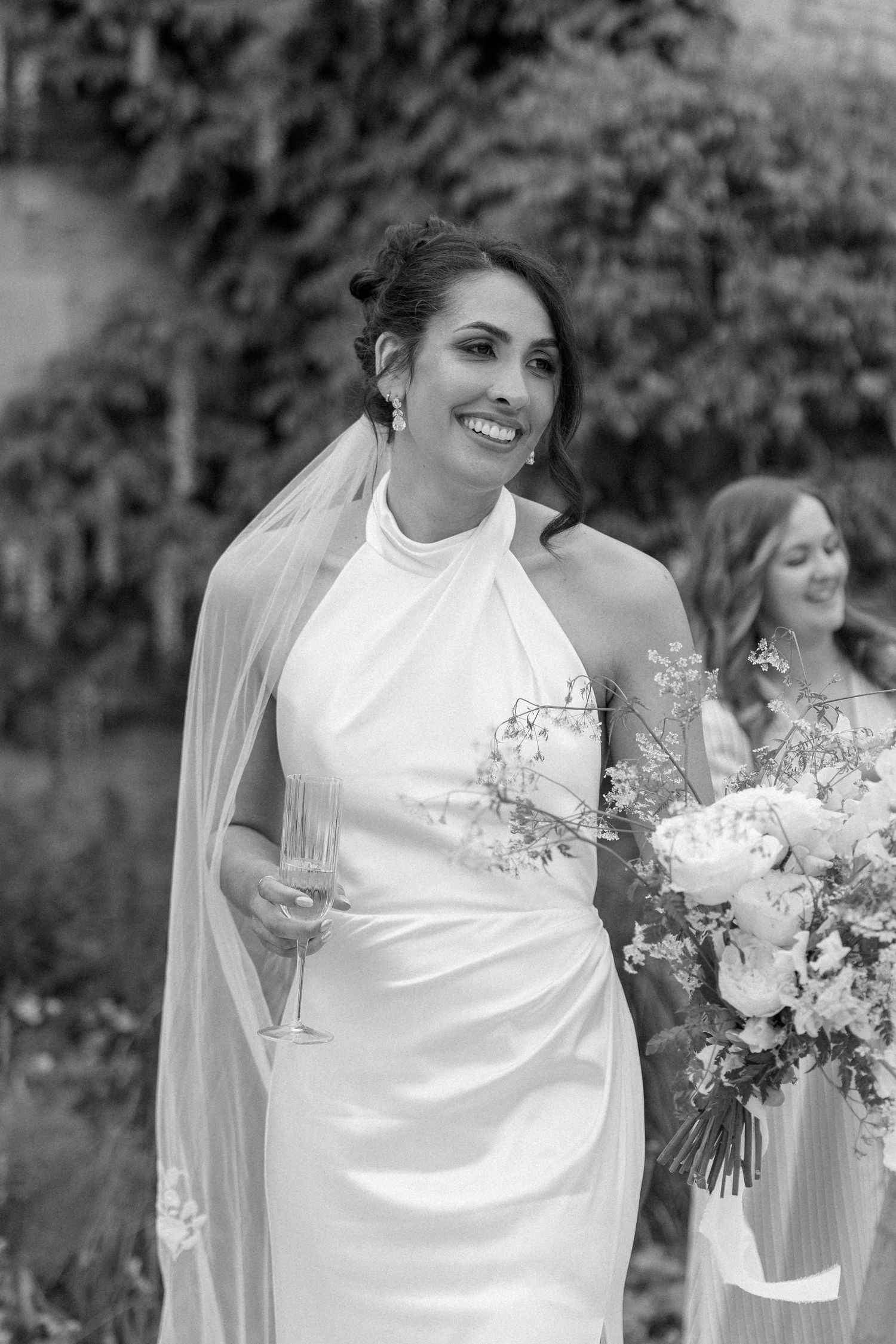 Elegant bride in halter neck satin gown holds white floral bouquet and champagne glass in black and white portrait.