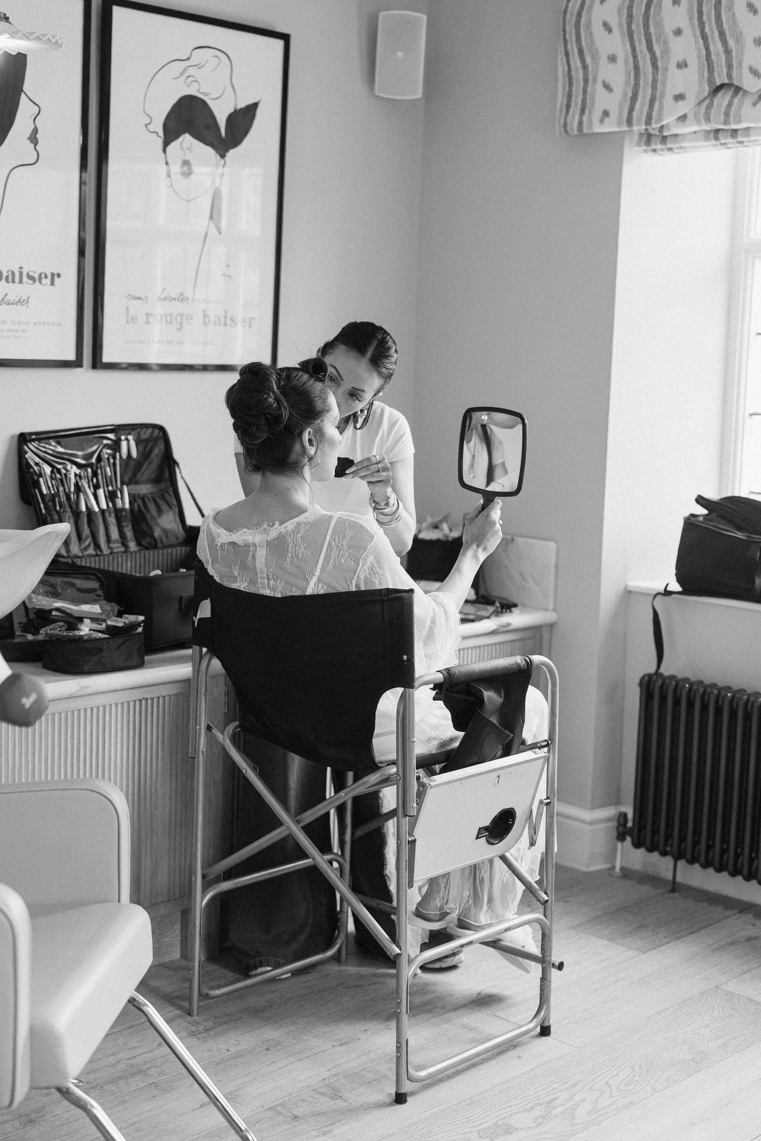 Black and white photo of a makeup artist applying makeup to a seated bride holding a mirror.