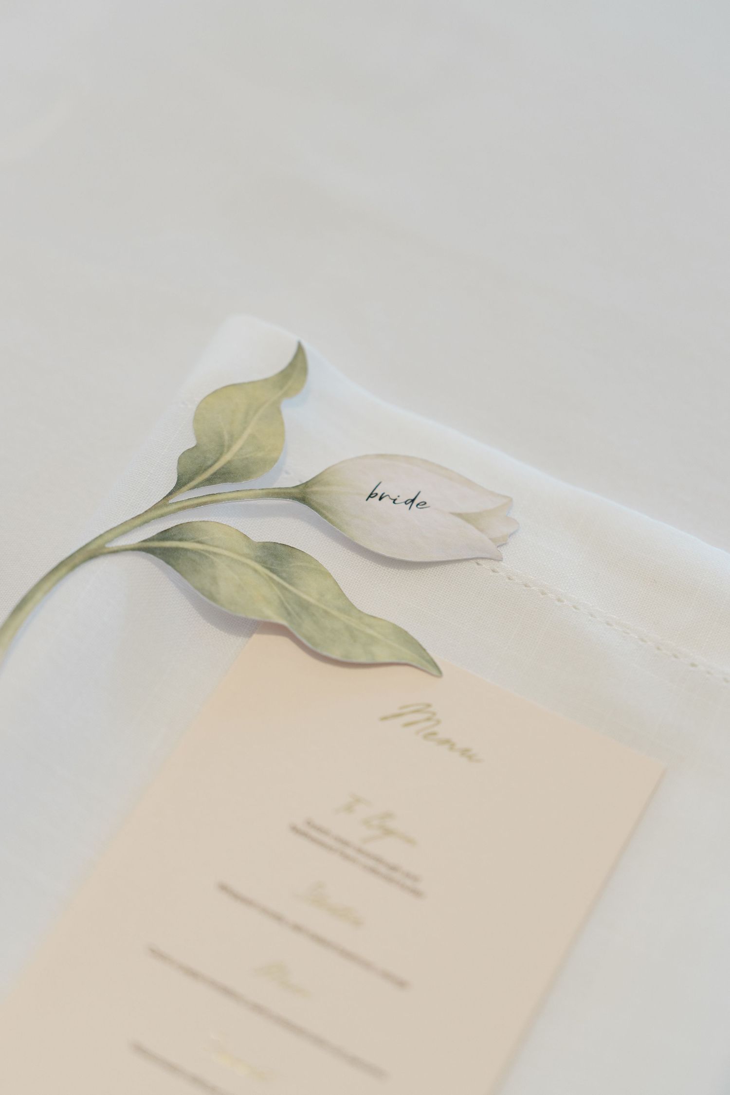 Elegant wedding place setting with tulip name card reading bride beside cream menu card on white linen.