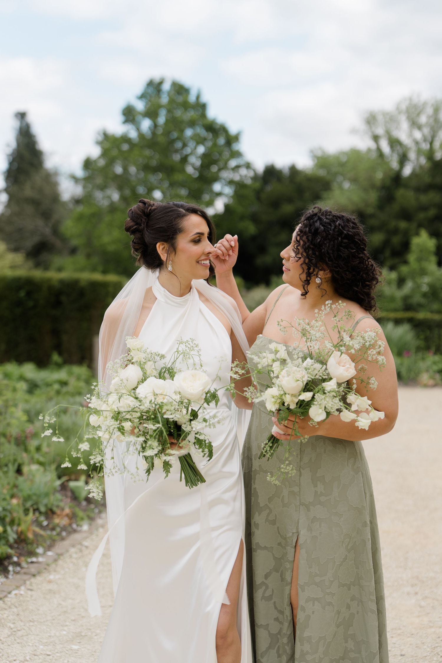 Bride in white halter gown and bridesmaid in sage green dress laugh while holding white bouquets outdoors.