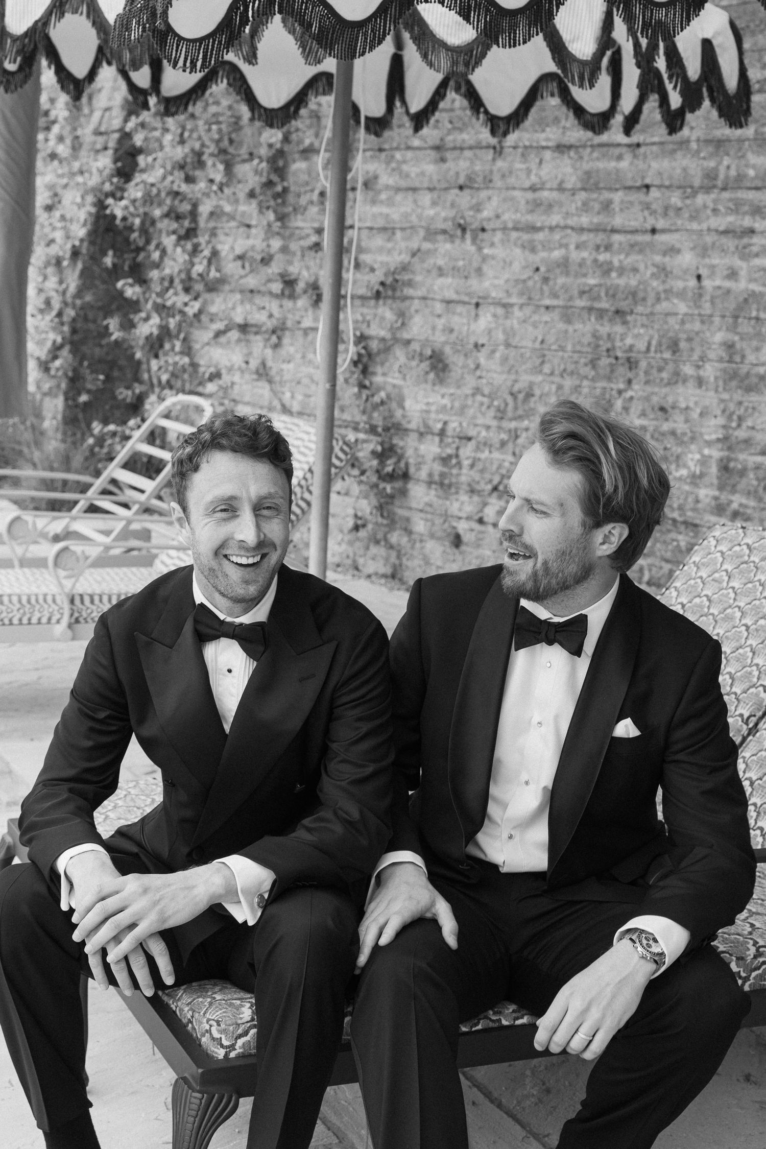 Two grooms in black tuxedos laugh together while seated outdoors at their wedding celebration.