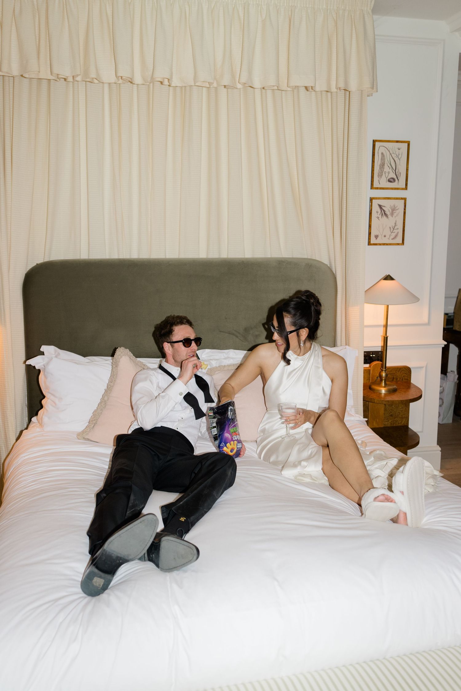 Newlyweds relax on white hotel bed in wedding attire, groom in sunglasses, bride in fluffy slippers.