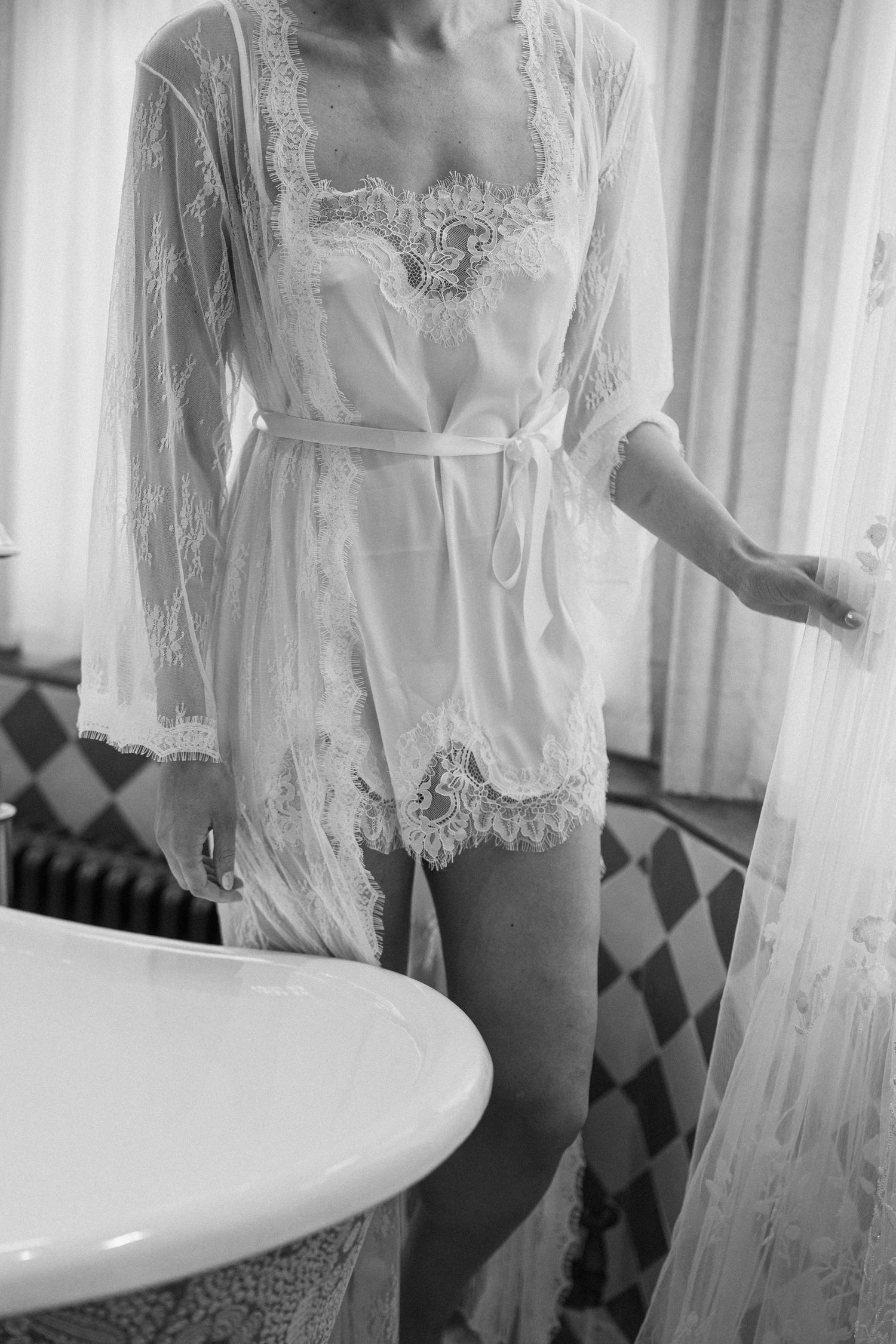 Bride in white lace robe standing by bathtub in black and white wedding preparation photo.