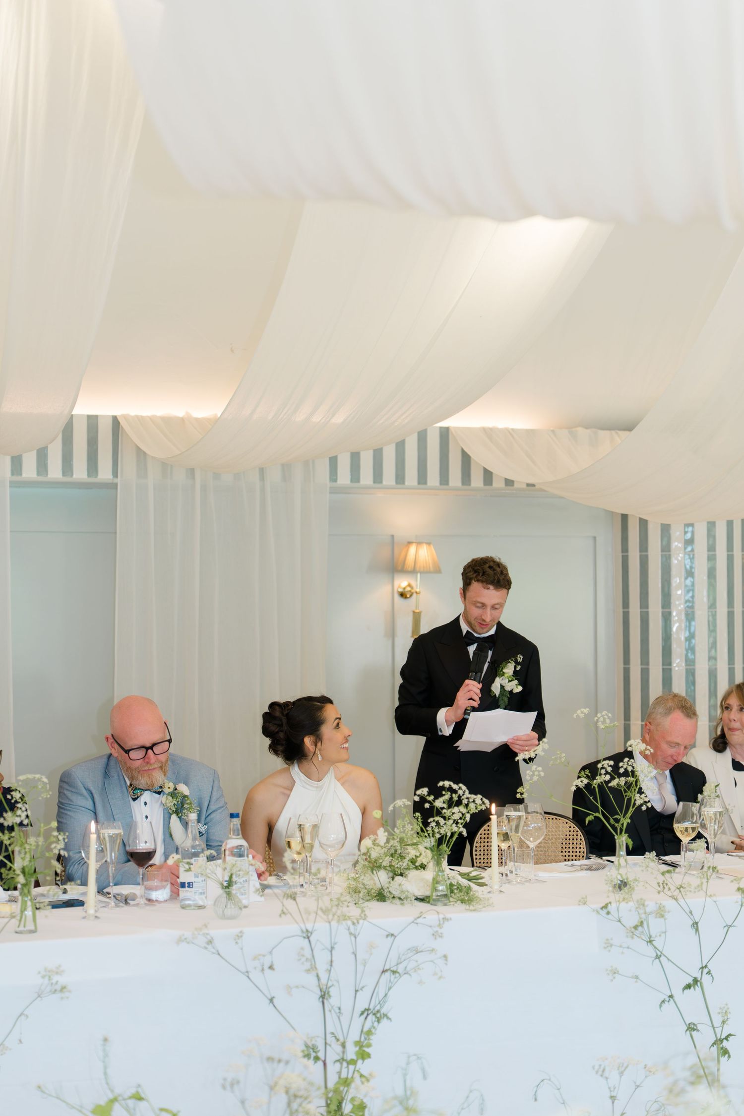 Groom delivers wedding speech at head table while bride smiles in a striped marquee reception venue.